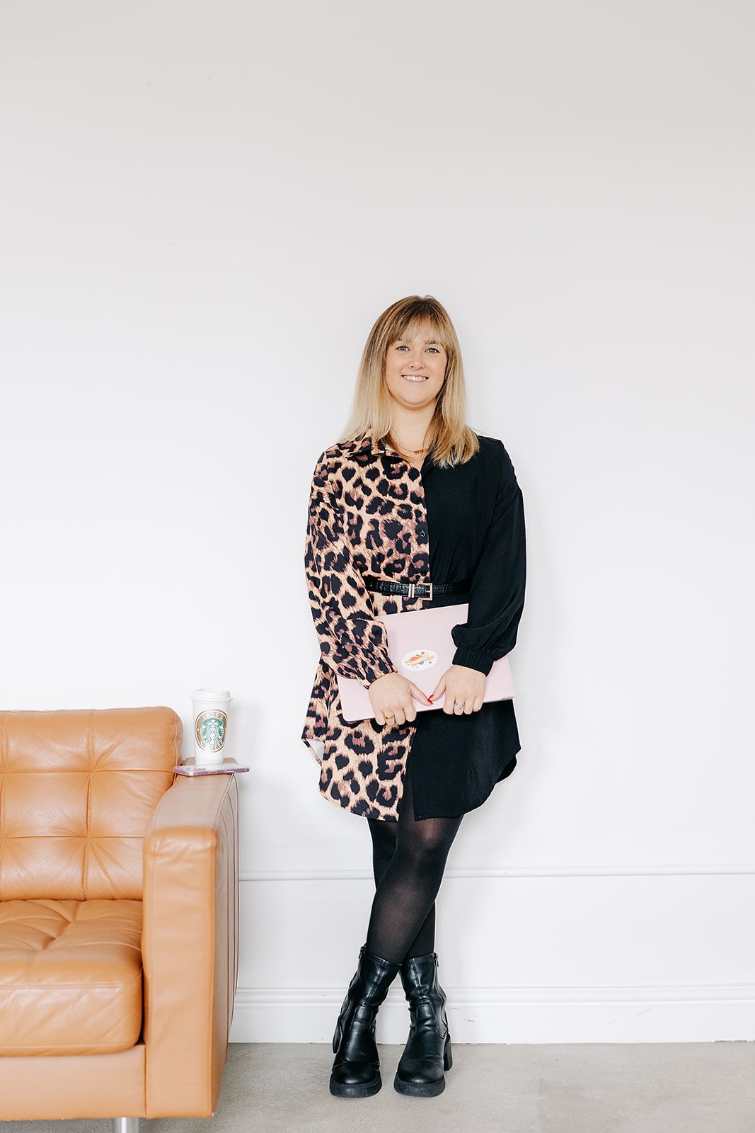 A person in a leopard print sweater and black boots sits on a tan leather couch in a minimalist white room.