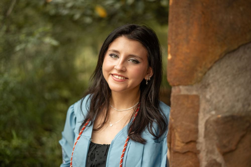 Person wearing a light blue jacket and black top smiles while standing outdoors near a stone wall.
