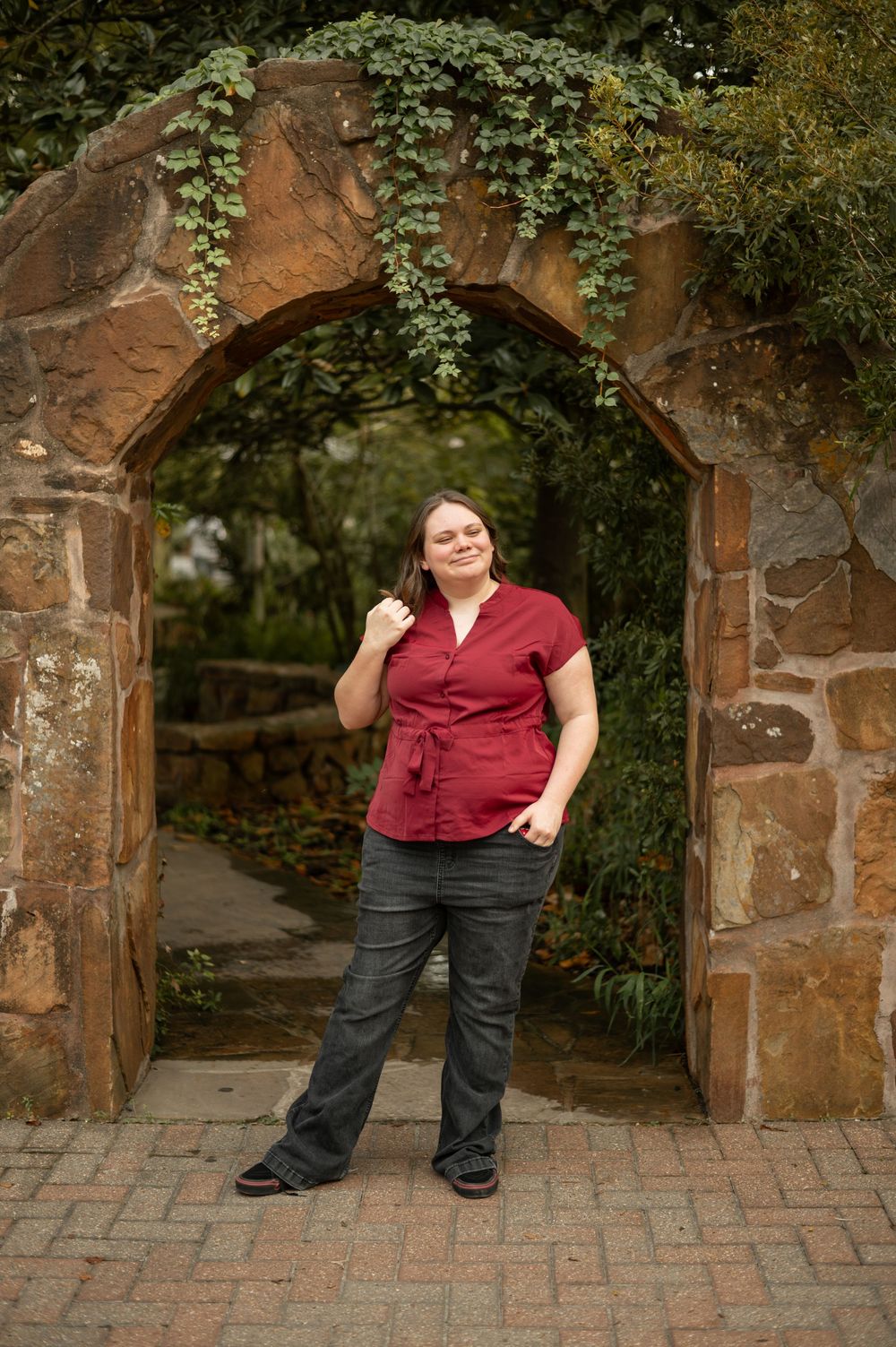 A person in a burgundy top and dark pants stands by a stone archway in a garden setting.