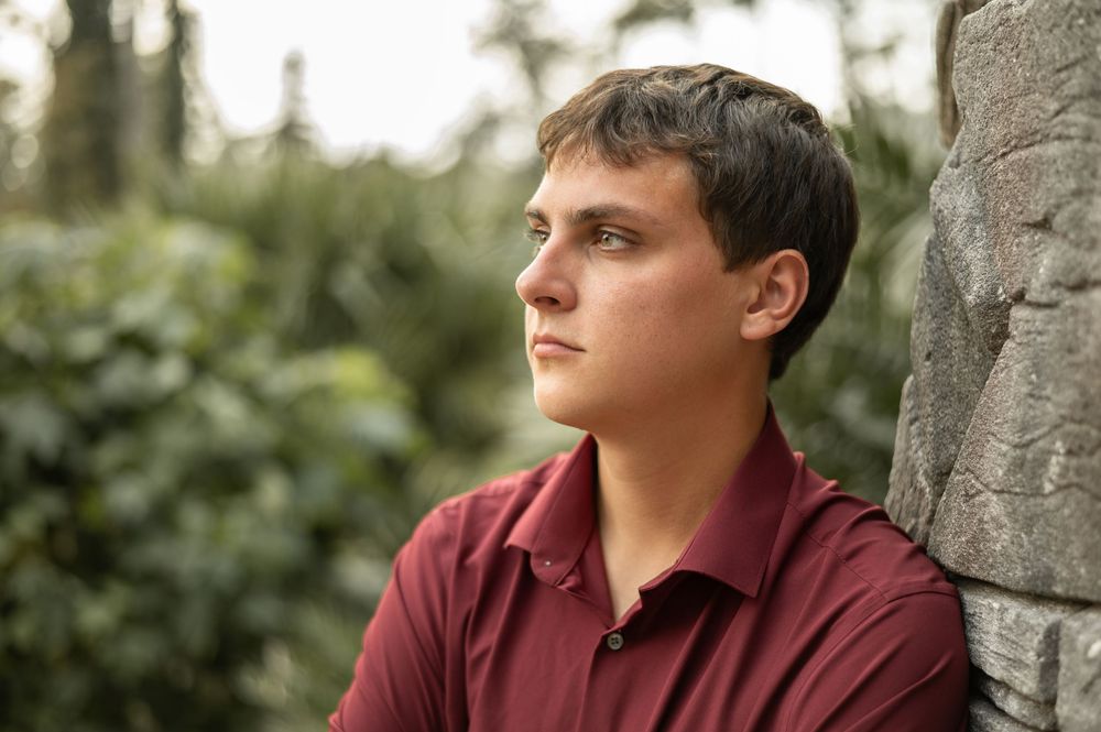 A person in a burgundy shirt leans against a stone wall while gazing thoughtfully into the distance.