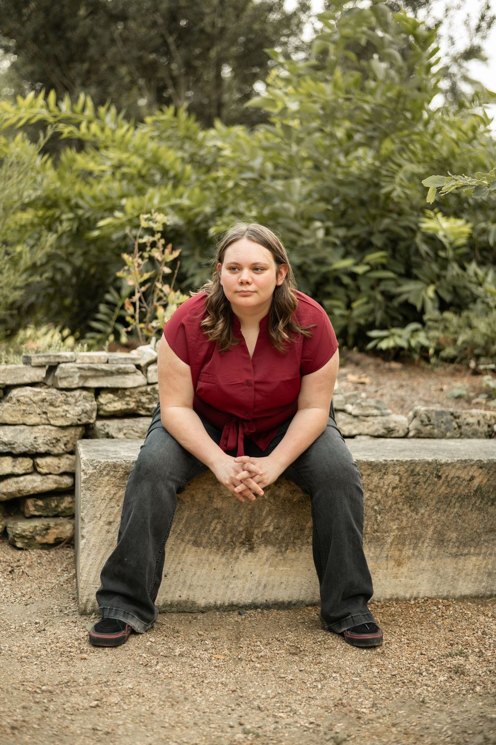 A person in a burgundy top and dark pants sits on a stone ledge in an outdoor garden setting.