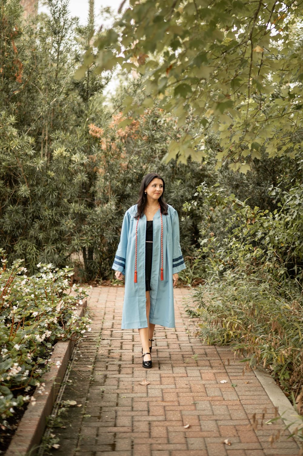 A person wearing a light blue kimono-style robe walks along a brick pathway surrounded by lush garden foliage.