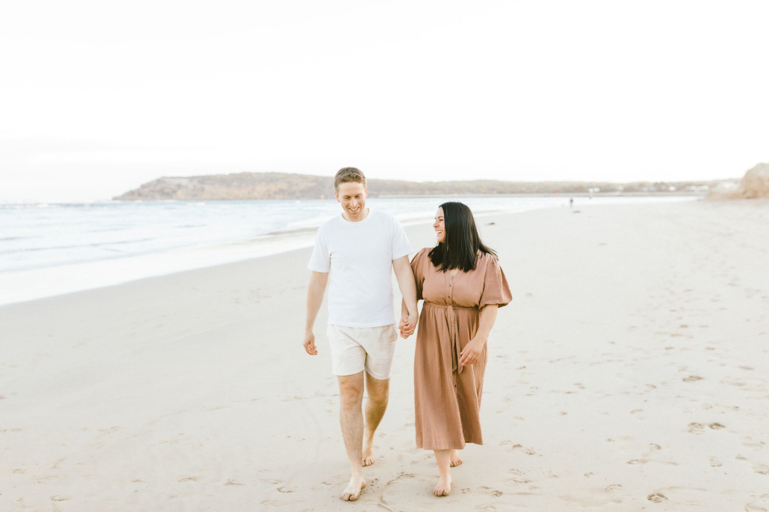 A couple walks along the sandy beach holding hands while wearing white and brown outfits.