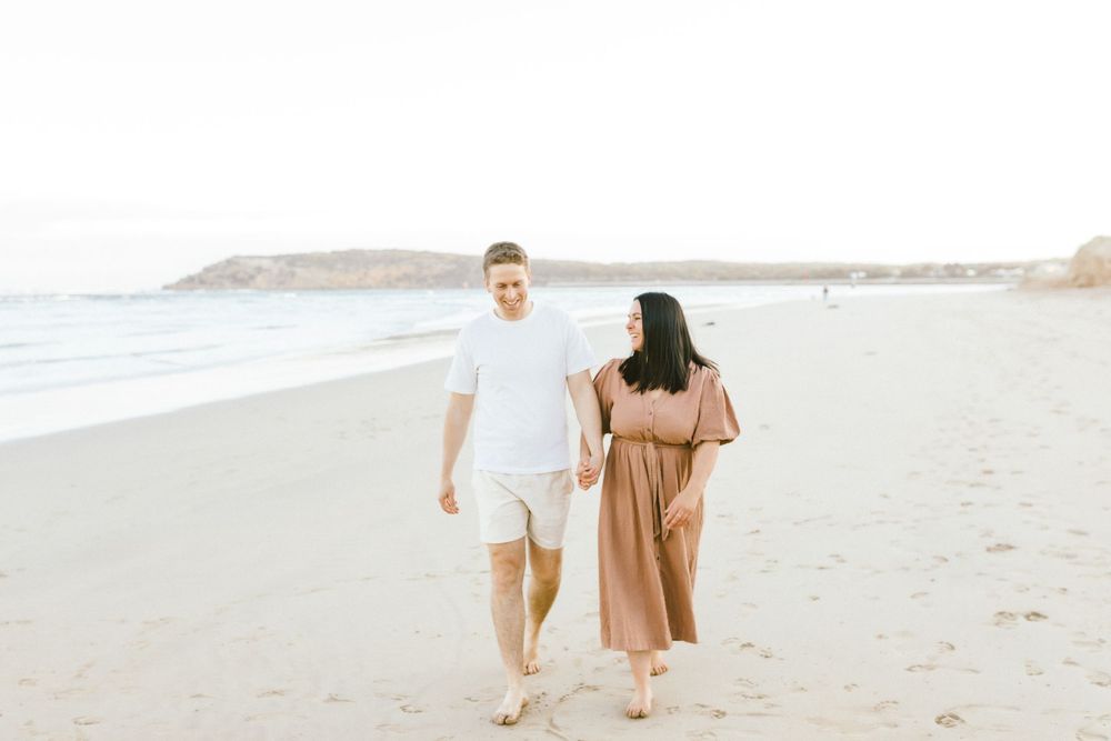 A couple walks along the sandy beach holding hands while wearing white and brown outfits.