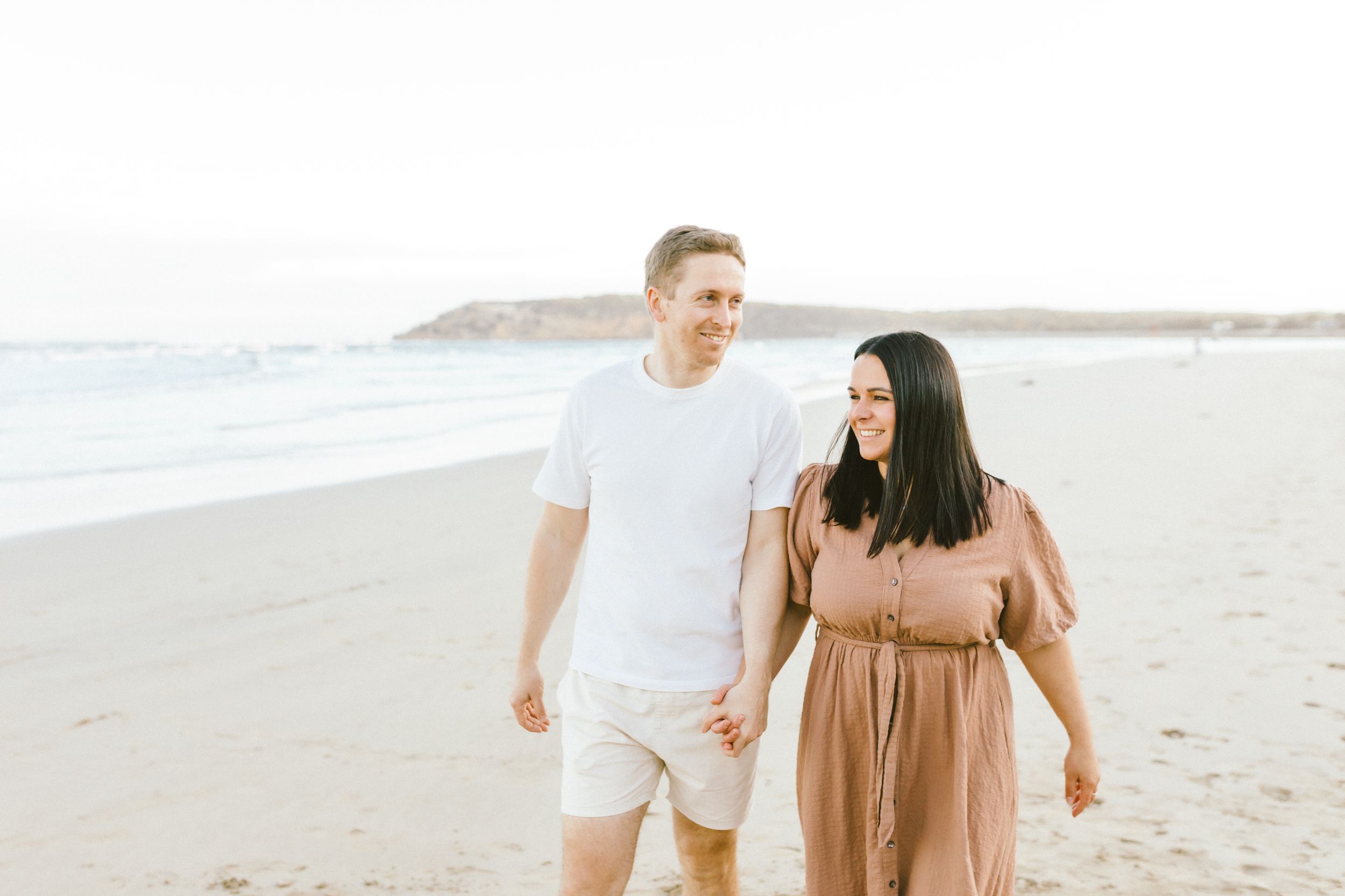 A couple walks together on a sandy beach during sunset while wearing white and brown outfits.