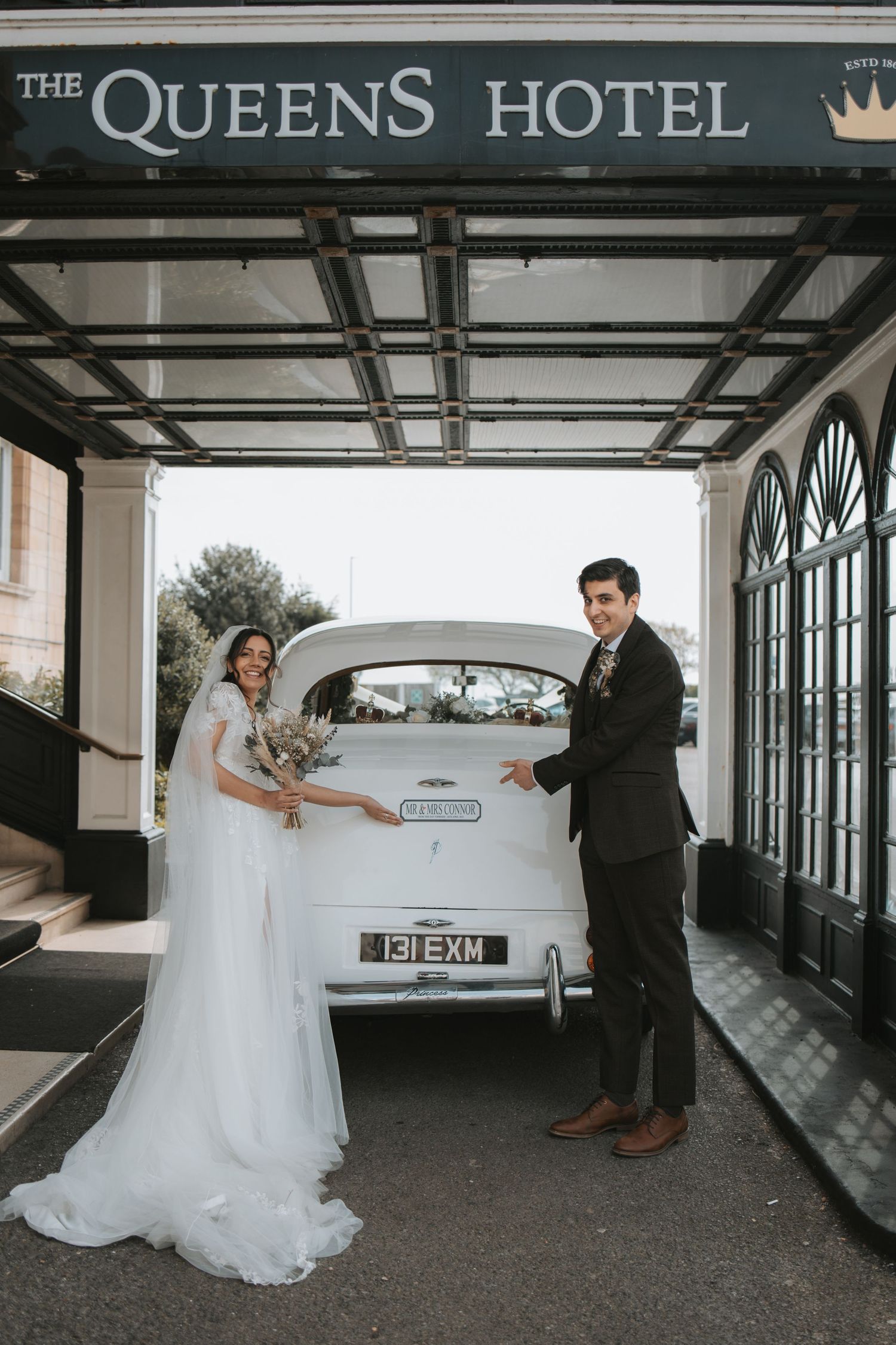 A wedding party poses outside The Queens Hotel with a vintage white car.