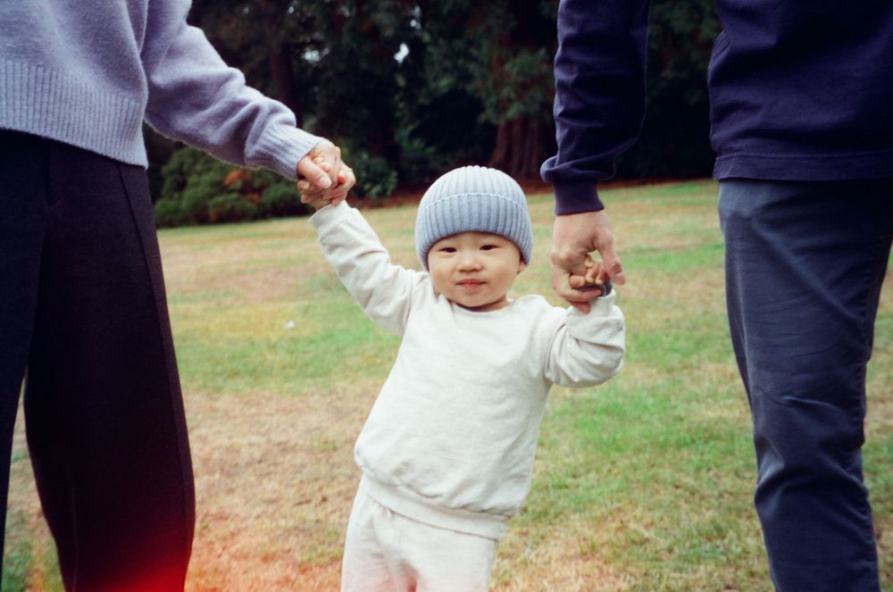 A small child in a white sweater and blue knit hat holds hands with adults while walking on grass.