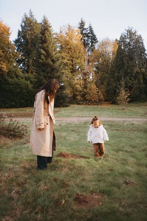 Person in a beige coat walks through a grassy field with autumn trees in the background on a sunny afternoon.