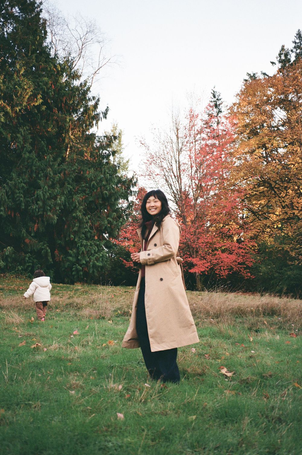 A person wearing a beige coat stands in a grassy field surrounded by colorful autumn trees on a cloudy day.