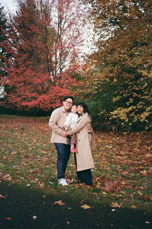 A couple embraces while standing on fallen autumn leaves with vibrant red and orange foliage in the background.