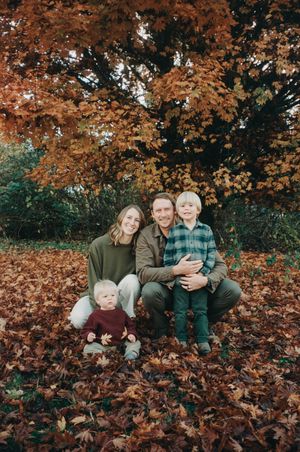 A family sits together on fallen autumn leaves beneath trees with vibrant orange foliage in a park setting.