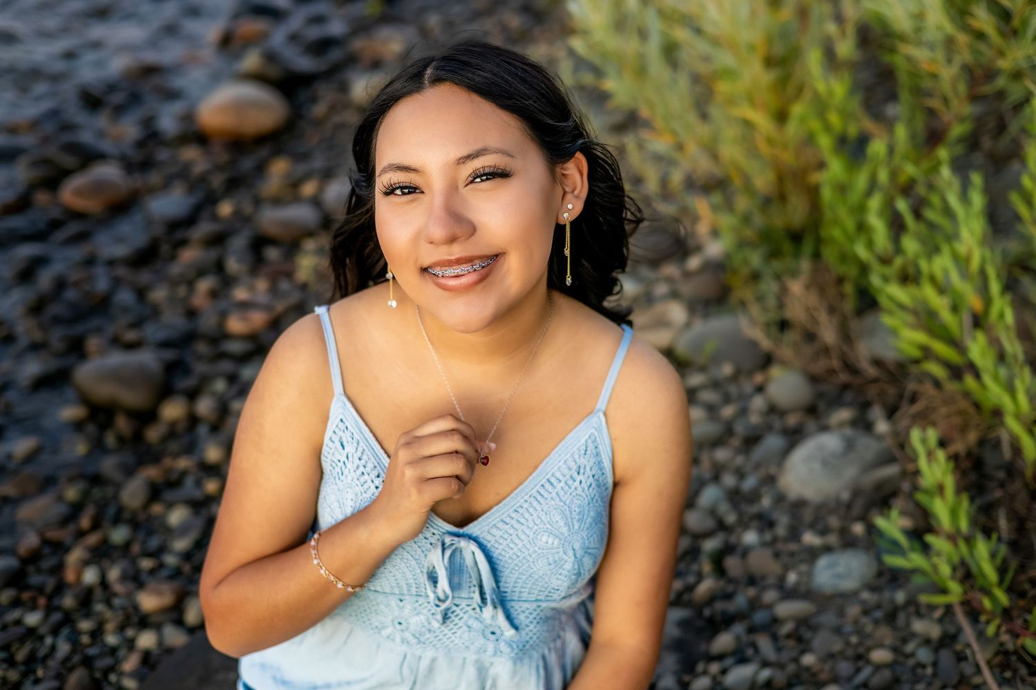 A person in a light blue dress sits on a rocky beach during an outdoor photo session.