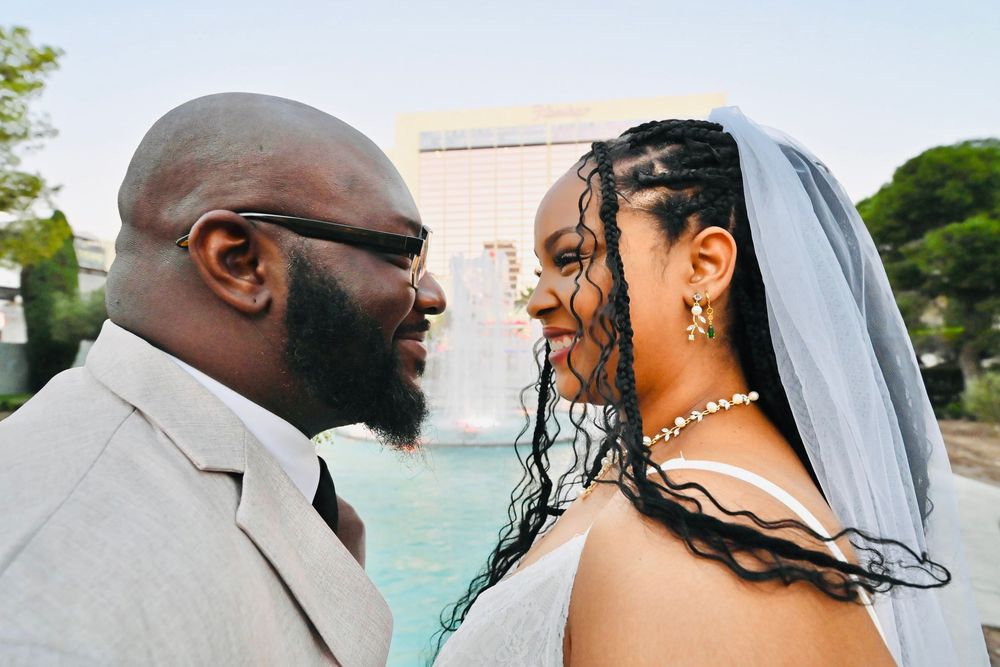 Newlyweds smile lovingly at each other outdoors, bride wearing a veil and groom in a gray suit.