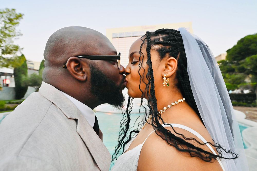 A bride and groom share a romantic kiss outdoors on their wedding day near a blue fountain.