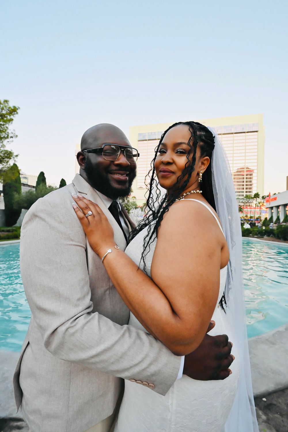 Smiling newlyweds embrace poolside in Las Vegas, bride in white gown with veil and groom in gray suit.