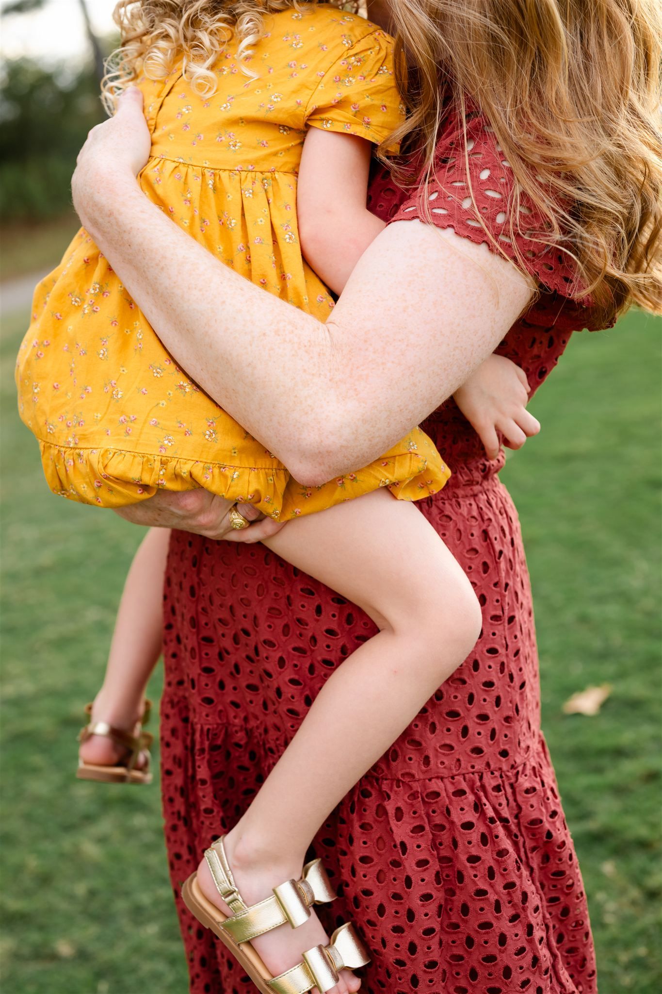 Close-up view of a mother in a burgundy dress holding a child wearing a mustard yellow outfit outdoors on grass.