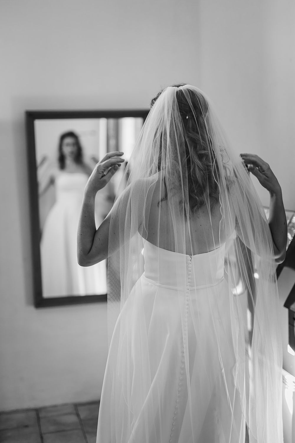 A bride adjusts her veil while looking at her reflection in a mirror during wedding preparations.