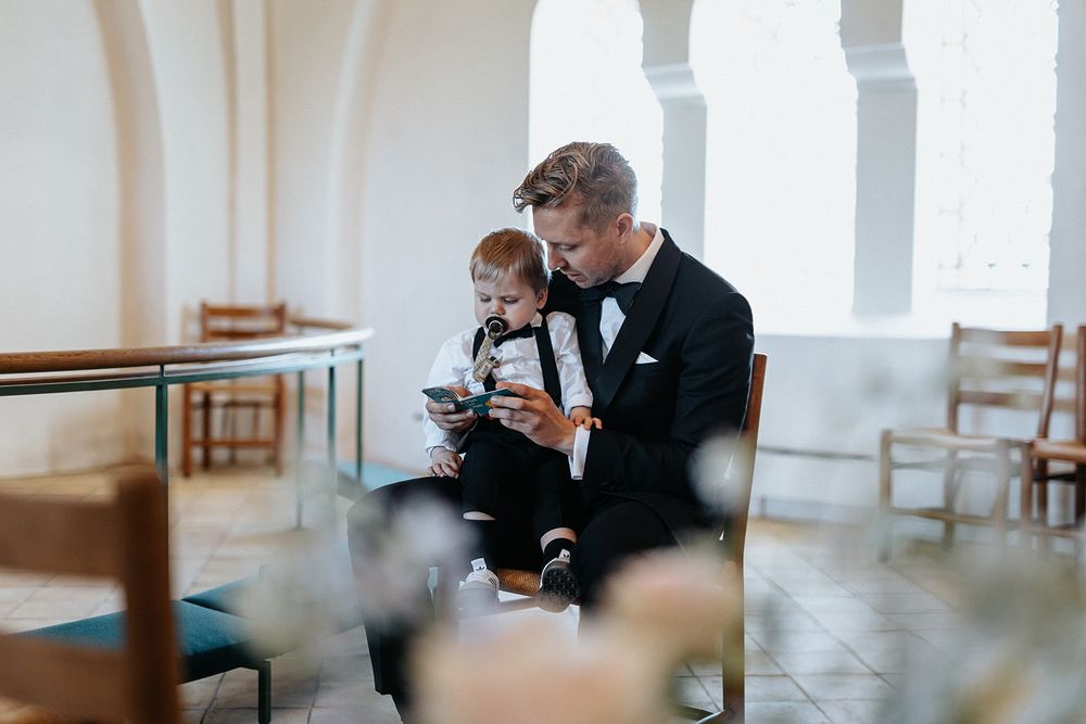 A groom in a black tuxedo sits with a small child looking at a device in a church setting.