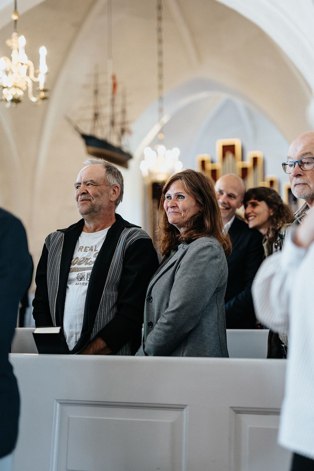 People seated in white church pews during a ceremony with elegant arched ceilings and chandeliers in the background.