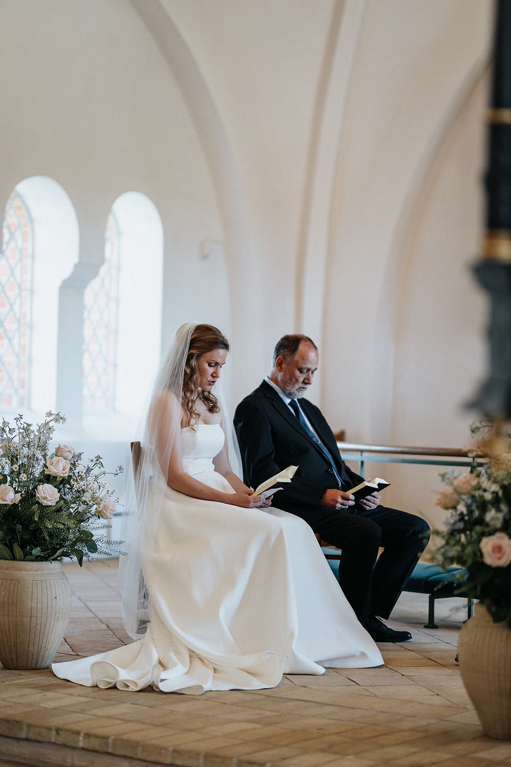 A wedding couple sits on a church bench reading together in a bright white chapel with arched windows.