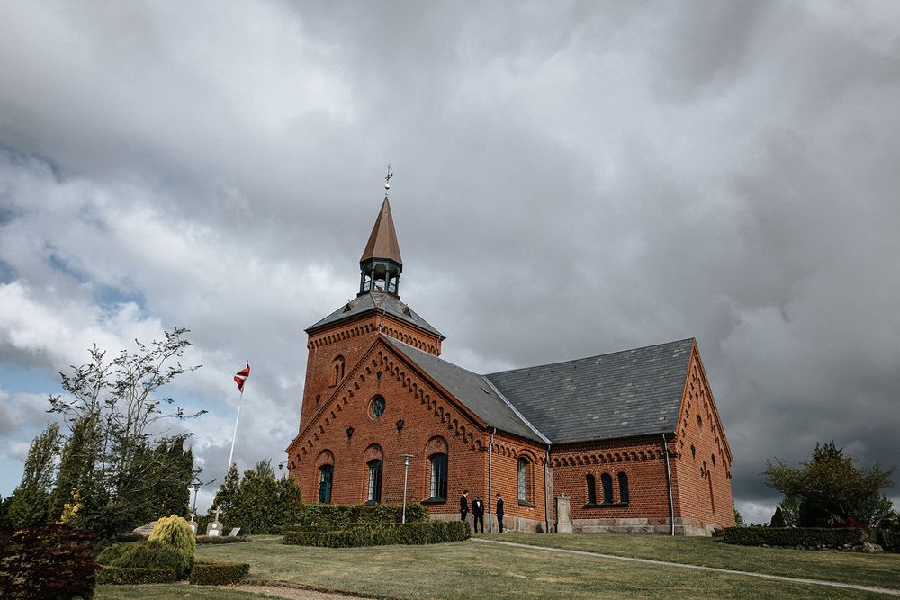 A dramatic panoramic view of a red brick church under stormy clouds with trees and buildings in the background.