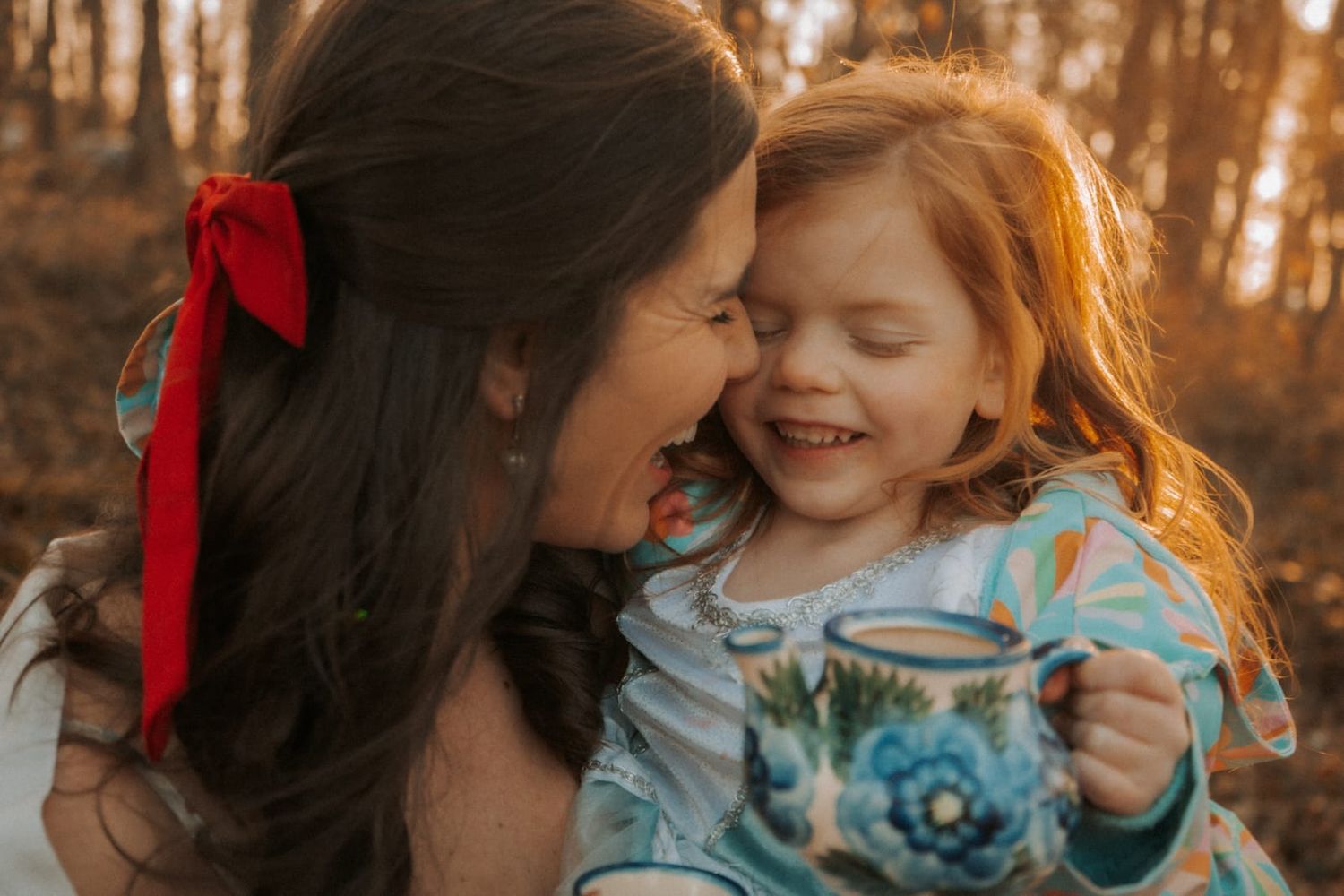 A smiling mother with a red bow kisses her laughing red-haired daughter holding a floral mug.
