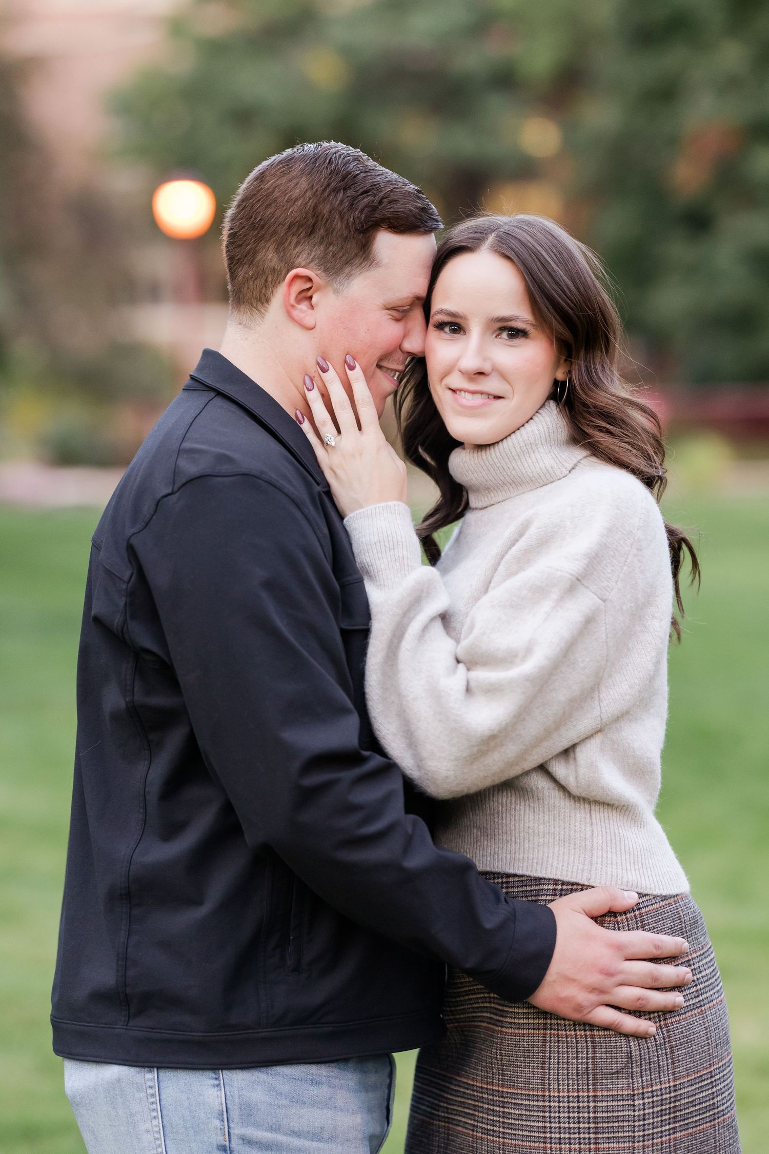 A couple shares an intimate moment outdoors, with her wearing a cream sweater and plaid skirt and him in a dark jacket.
