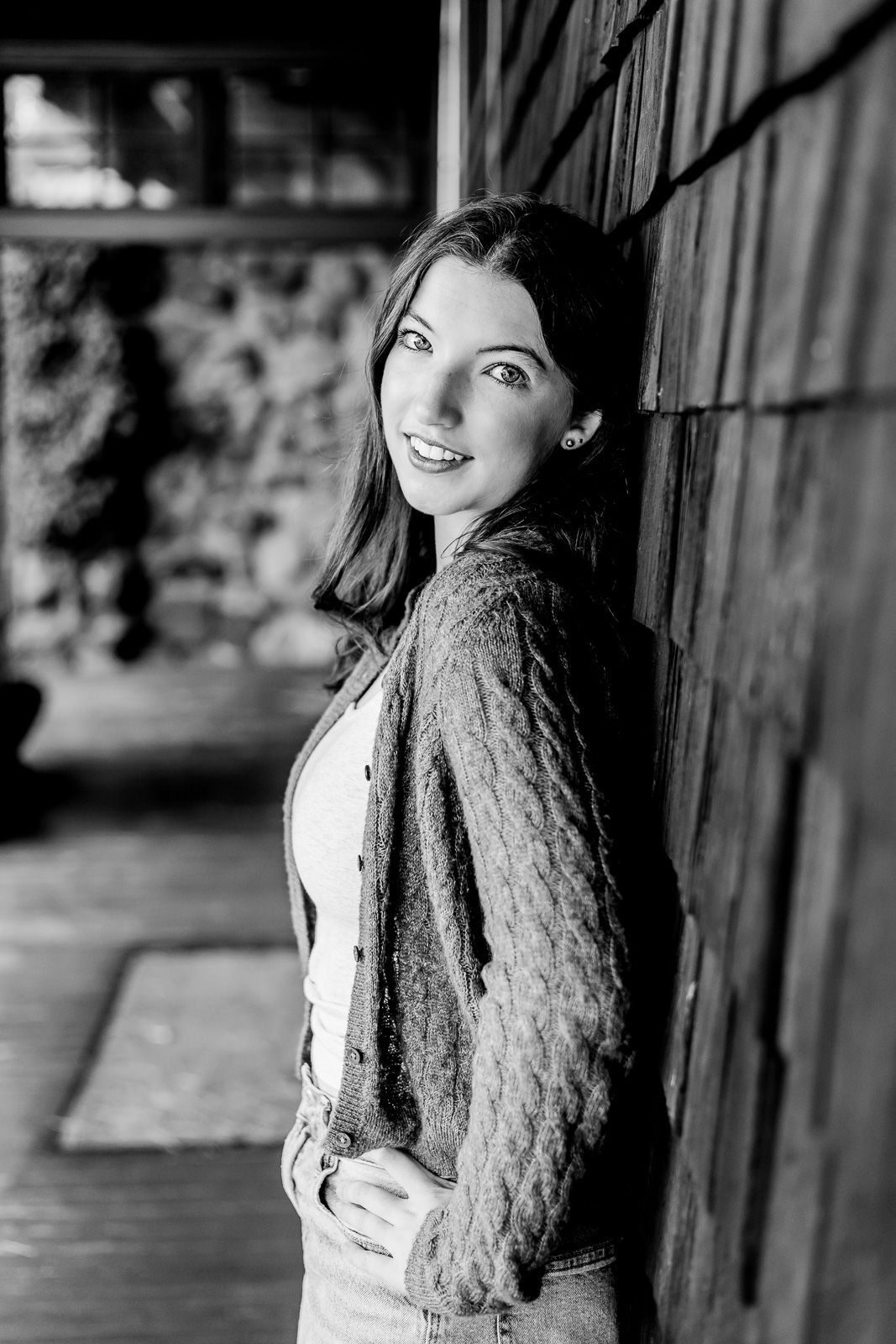 Black and white portrait of someone in a knit cardigan leaning against a wooden wall with a bright smile.