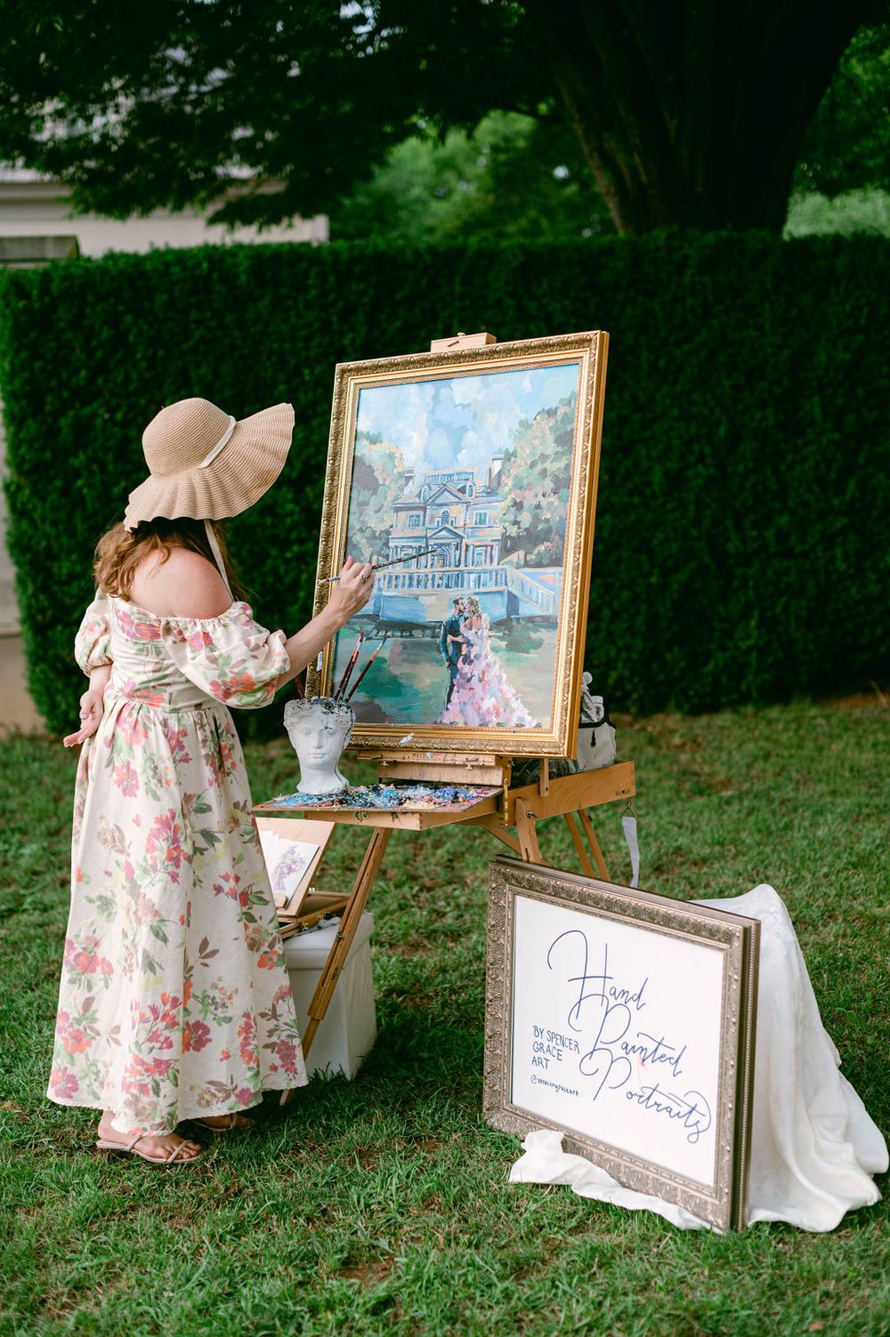 Artist in floral dress and sunhat painting at an easel in front of a lush garden hedge.
