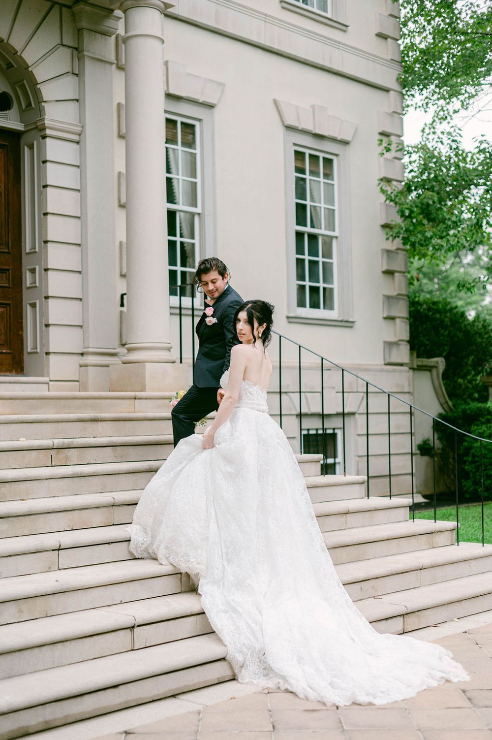 Elegant wedding couple poses on stone steps of historic white mansion.
