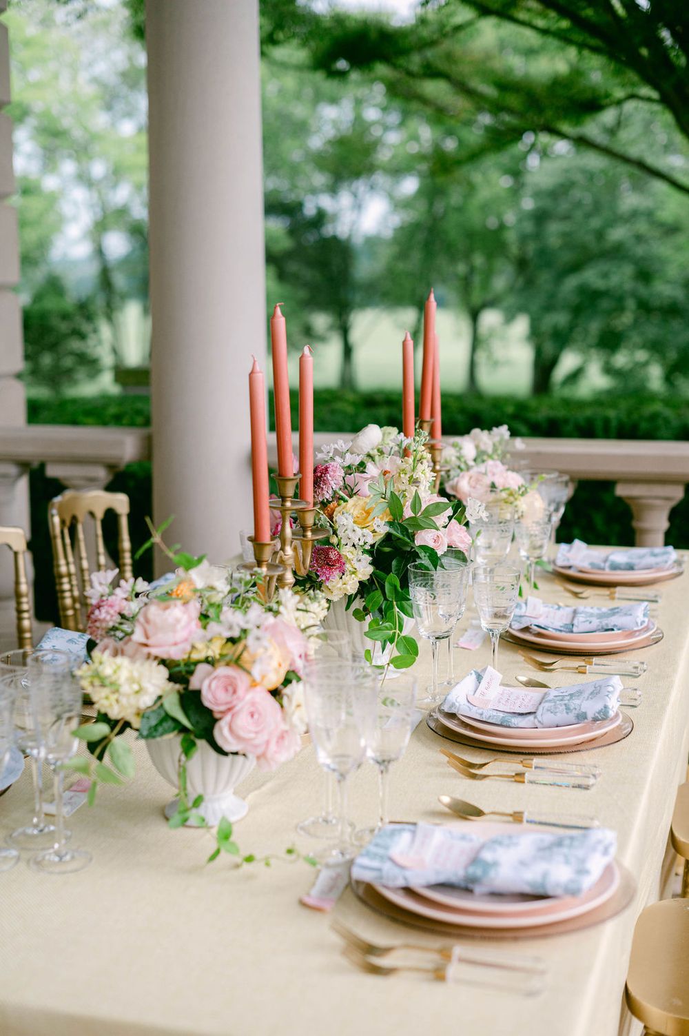Wedding reception table decorated with pink floral arrangements, coral candles and delicate place settings under a covered porch.