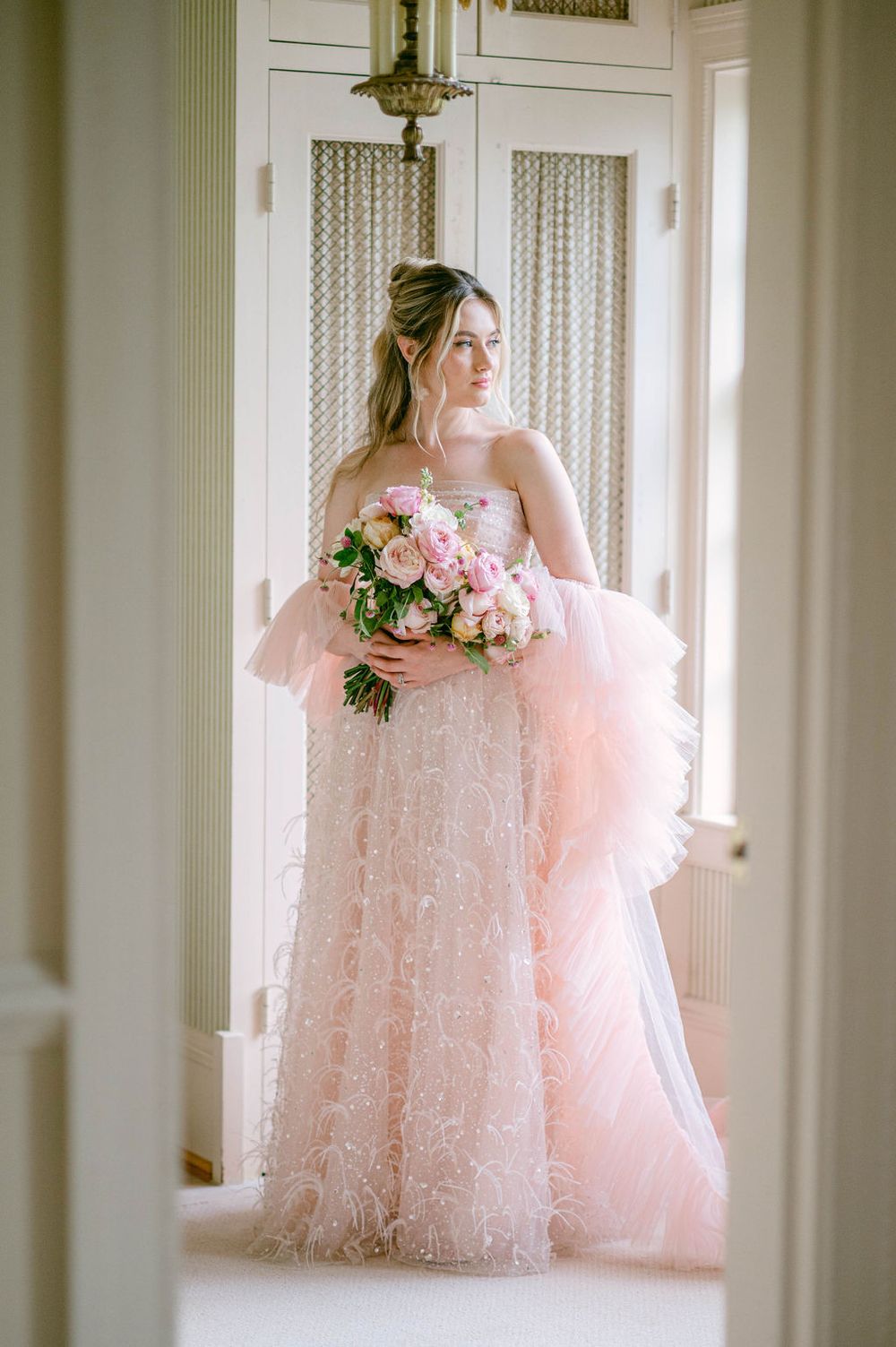 Ethereal pink tulle ballgown dress with lace details photographed in a bright white interior hallway.