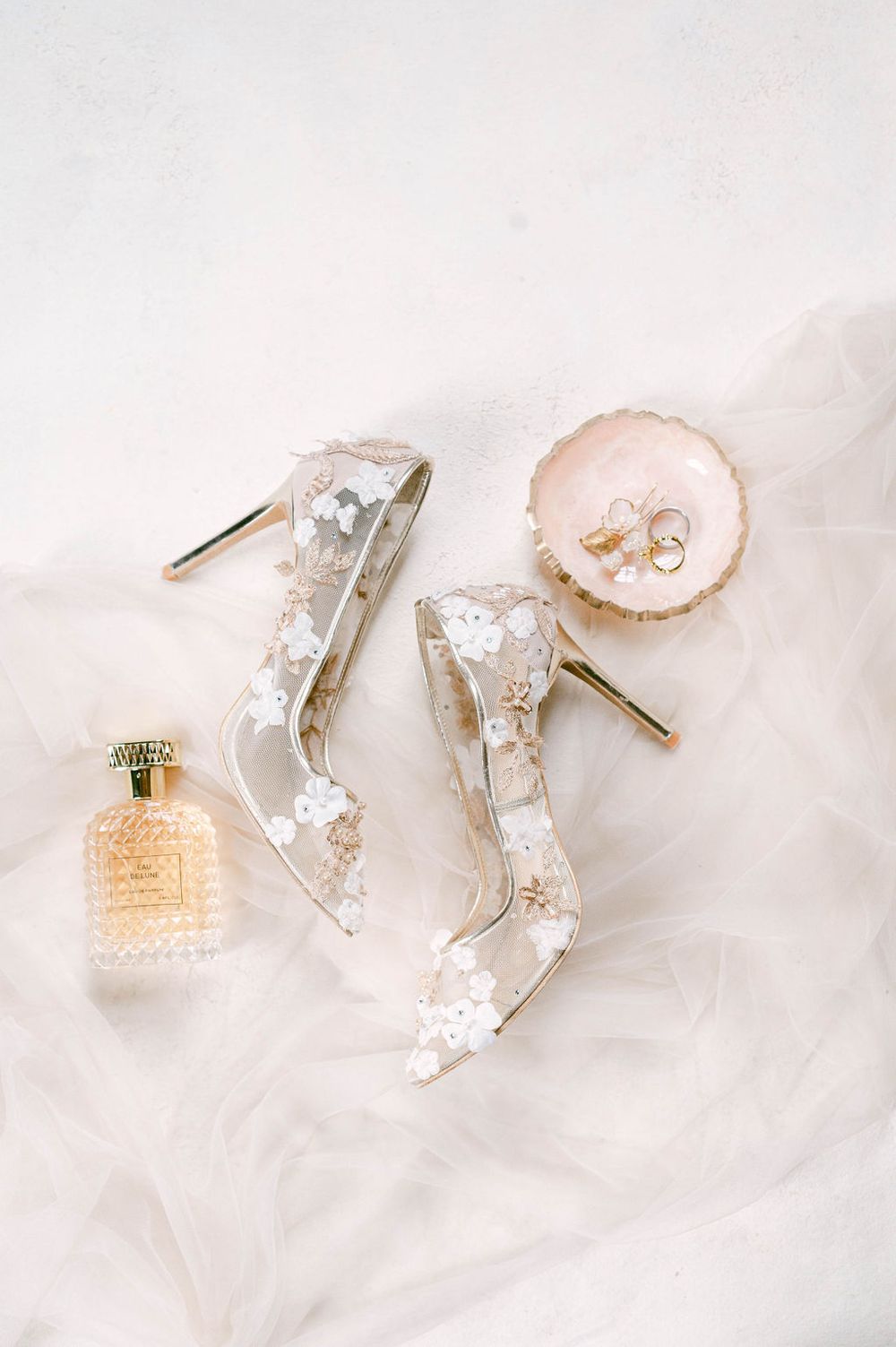 Delicate bridal accessories including clear heels, perfume bottle and jewelry on white fabric.
