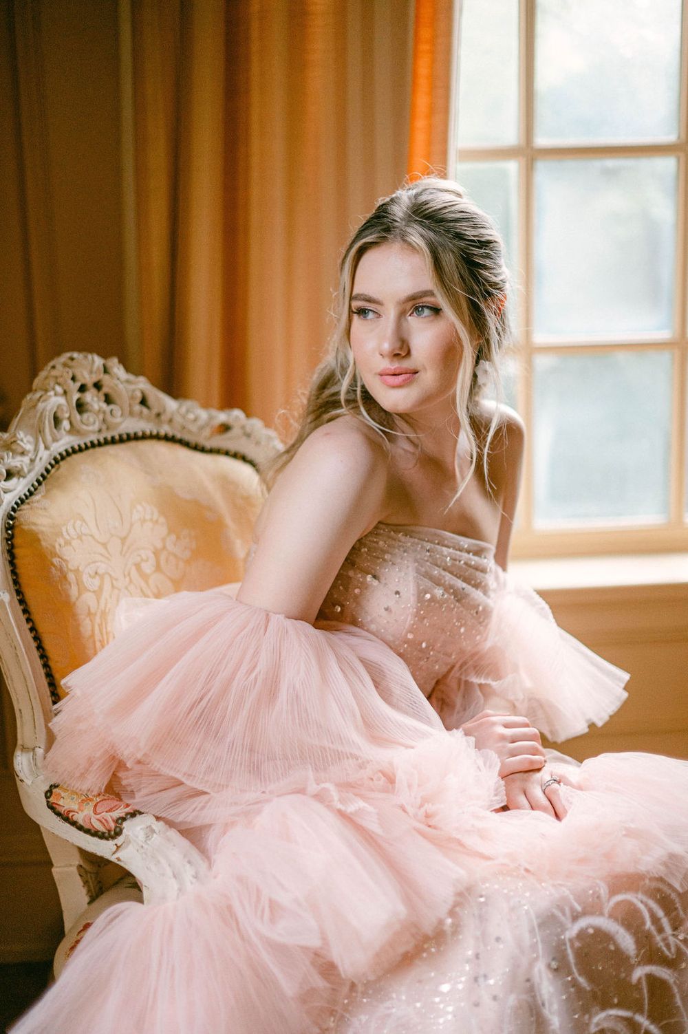 Elegant pink tulle gown displayed on an ornate vintage chair in a sunlit room.