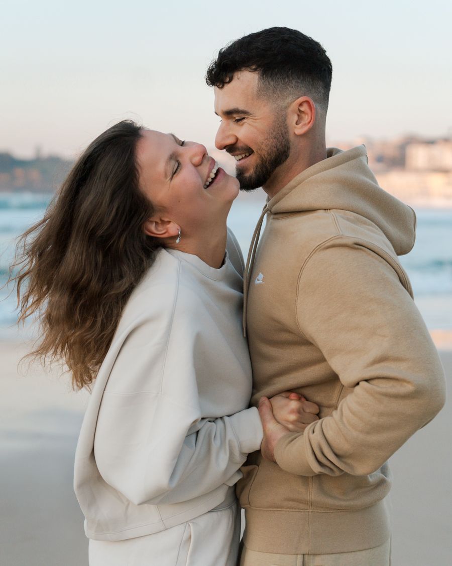 Couple sharing joyful moments and laughter at Bondi beach during sunrise while wearing cozy sweaters. Photoshoot in Sydney with a couple
