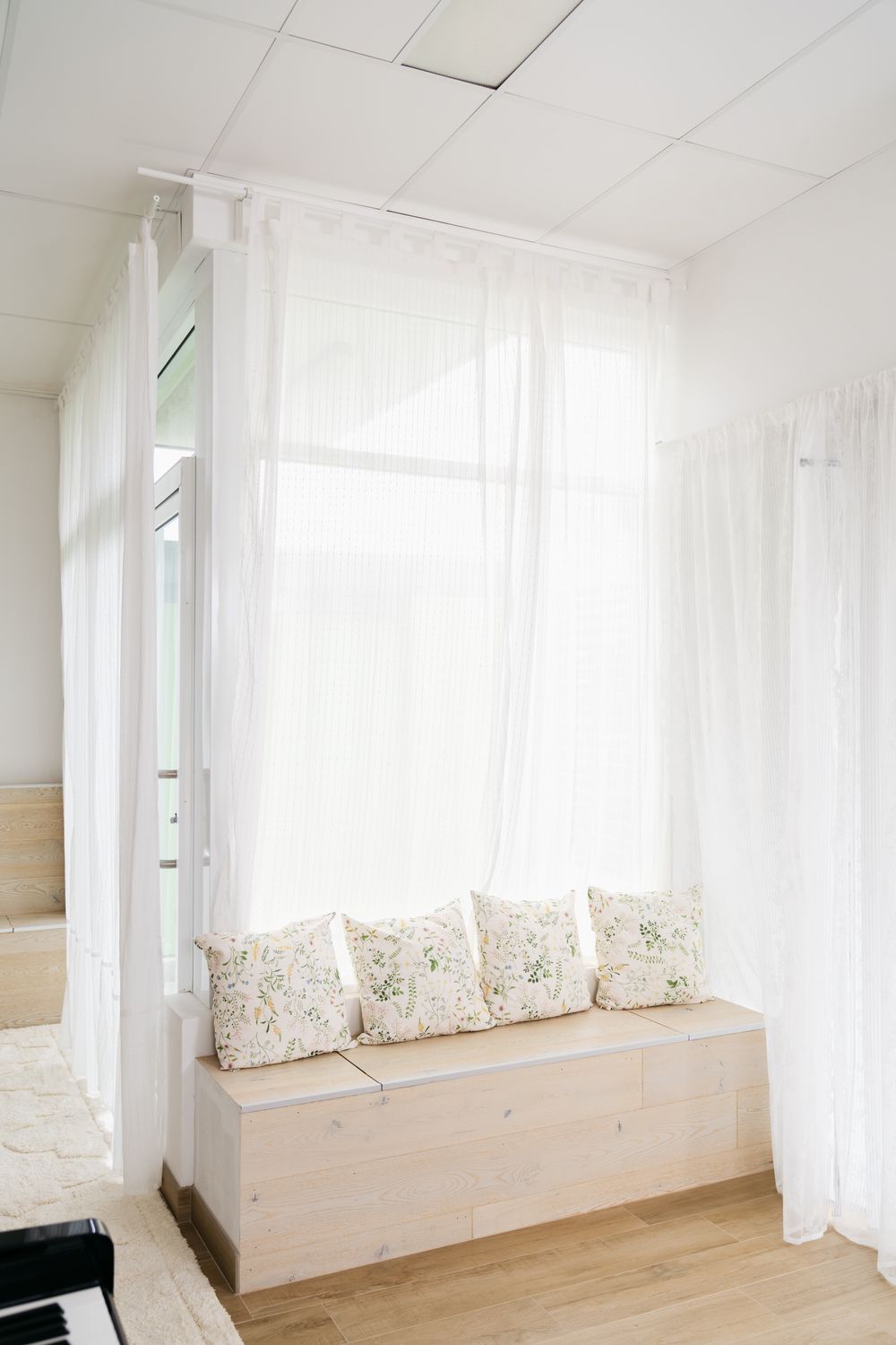 Bright white window nook with floral cushions on wooden bench and sheer curtains.