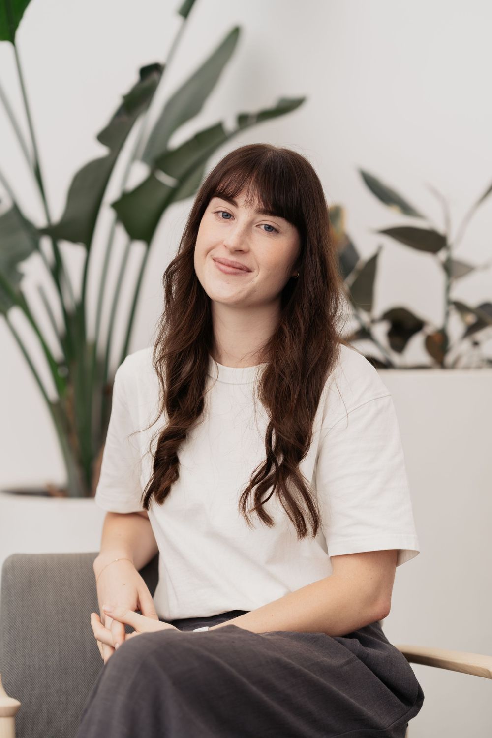 A person in a white t-shirt sits casually against a backdrop of indoor plants.