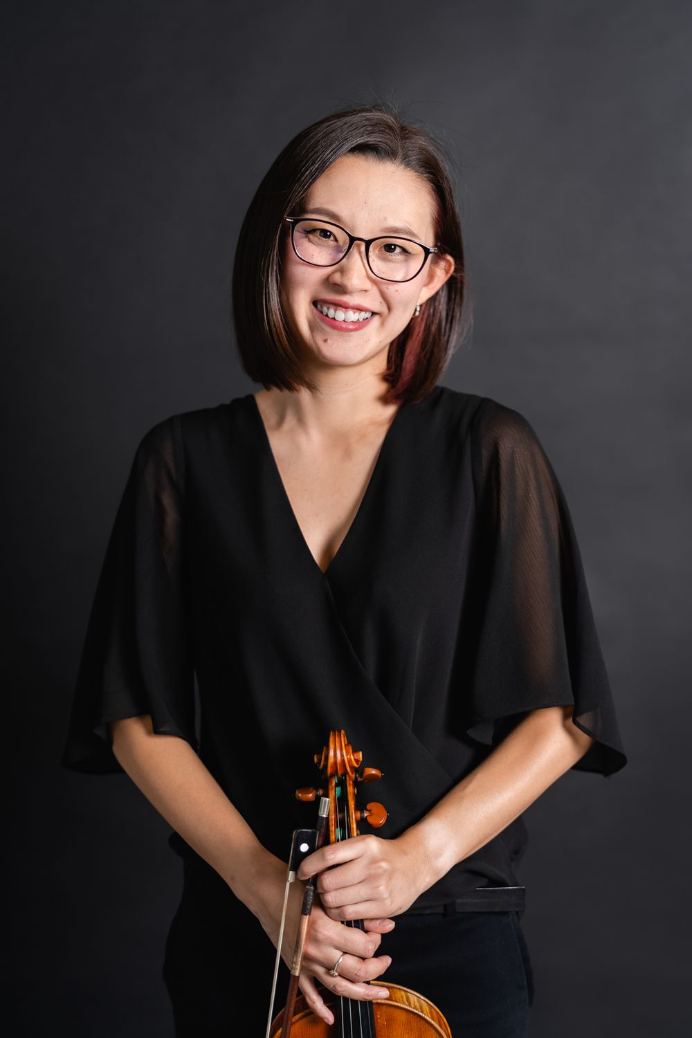 Professional musician poses with violin against dark background in elegant black attire.