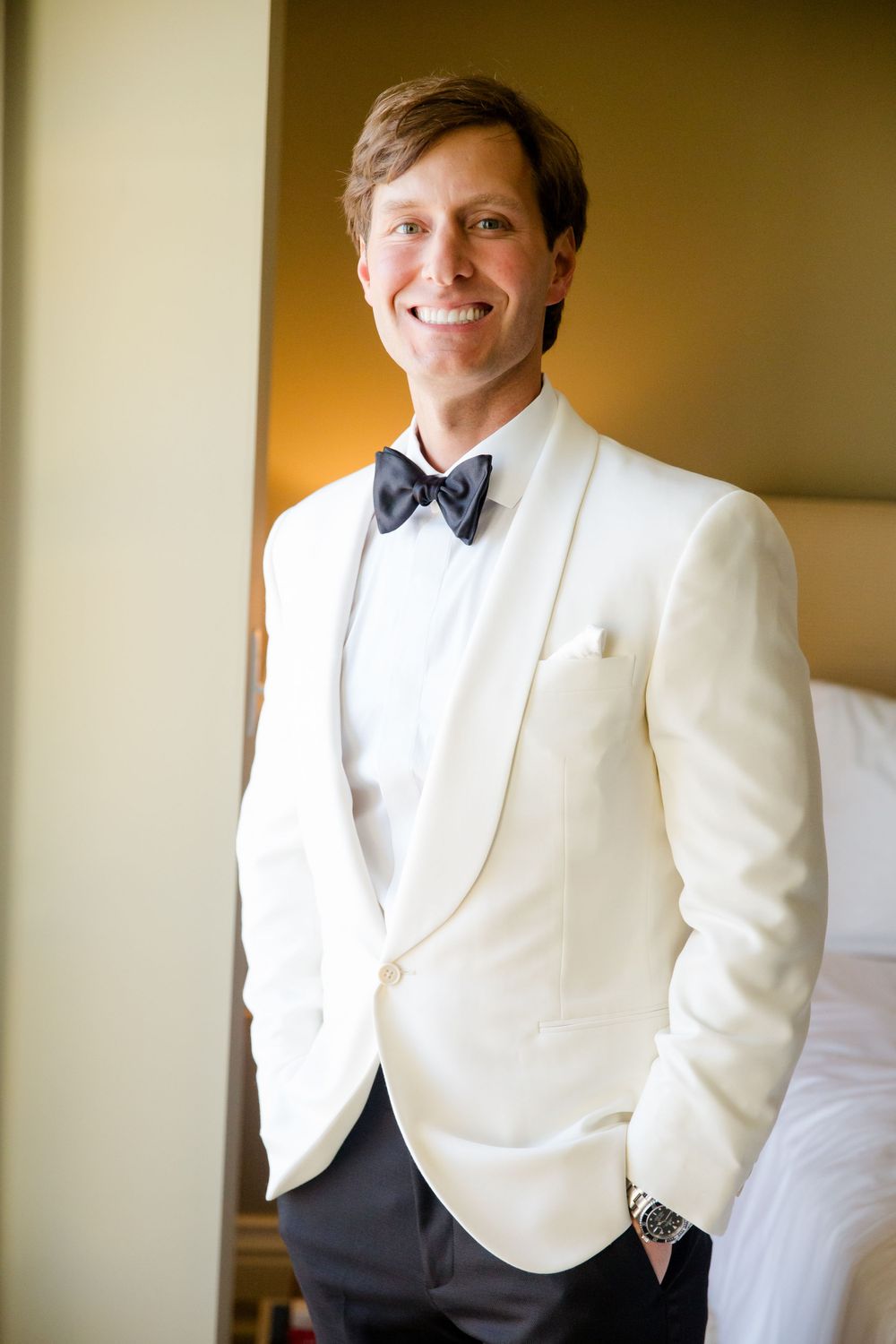 A groom in a white dinner jacket and bow tie smiles while getting ready for his wedding.