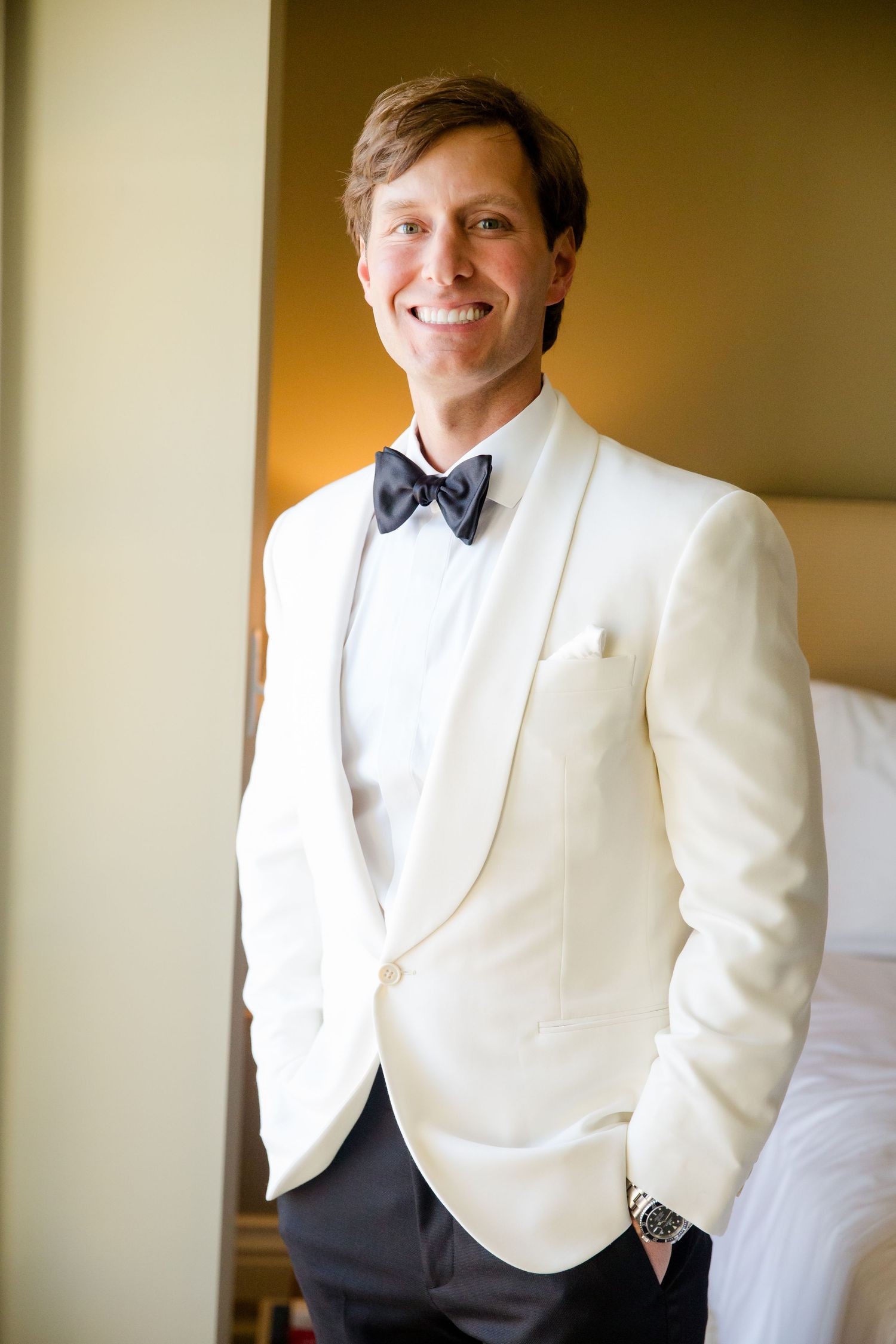 A groom in a white dinner jacket and bow tie smiles while getting ready for his wedding.