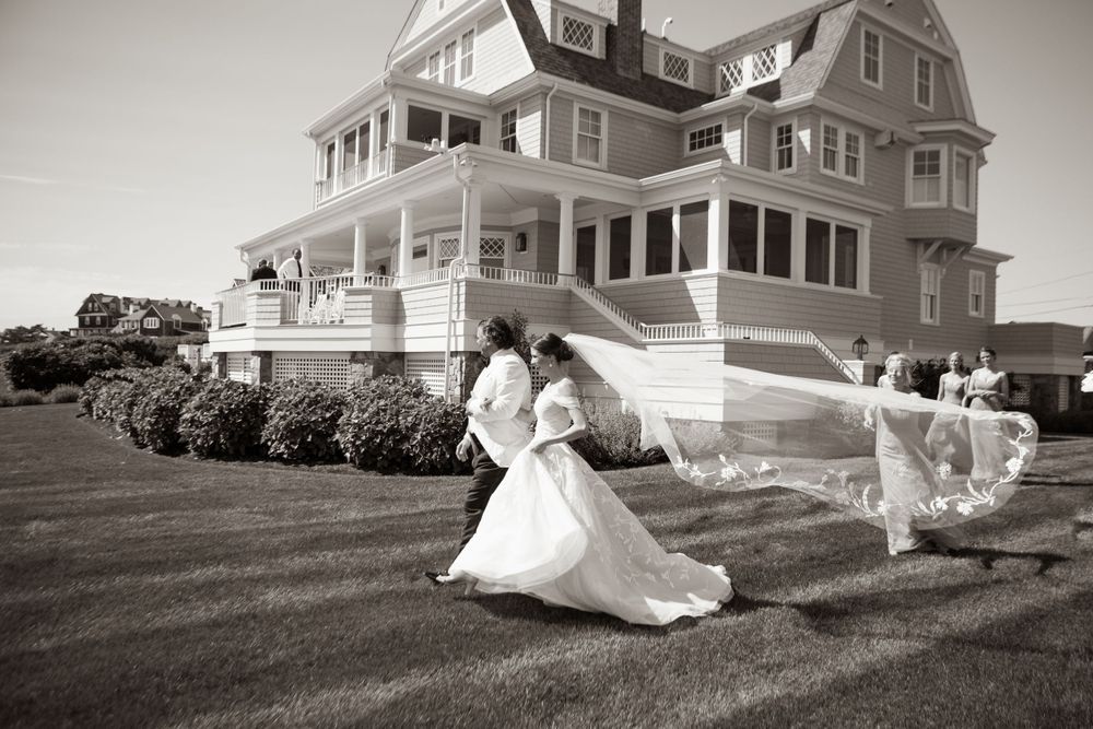 A black and white photo of newlyweds running on the lawn of a grand coastal estate.