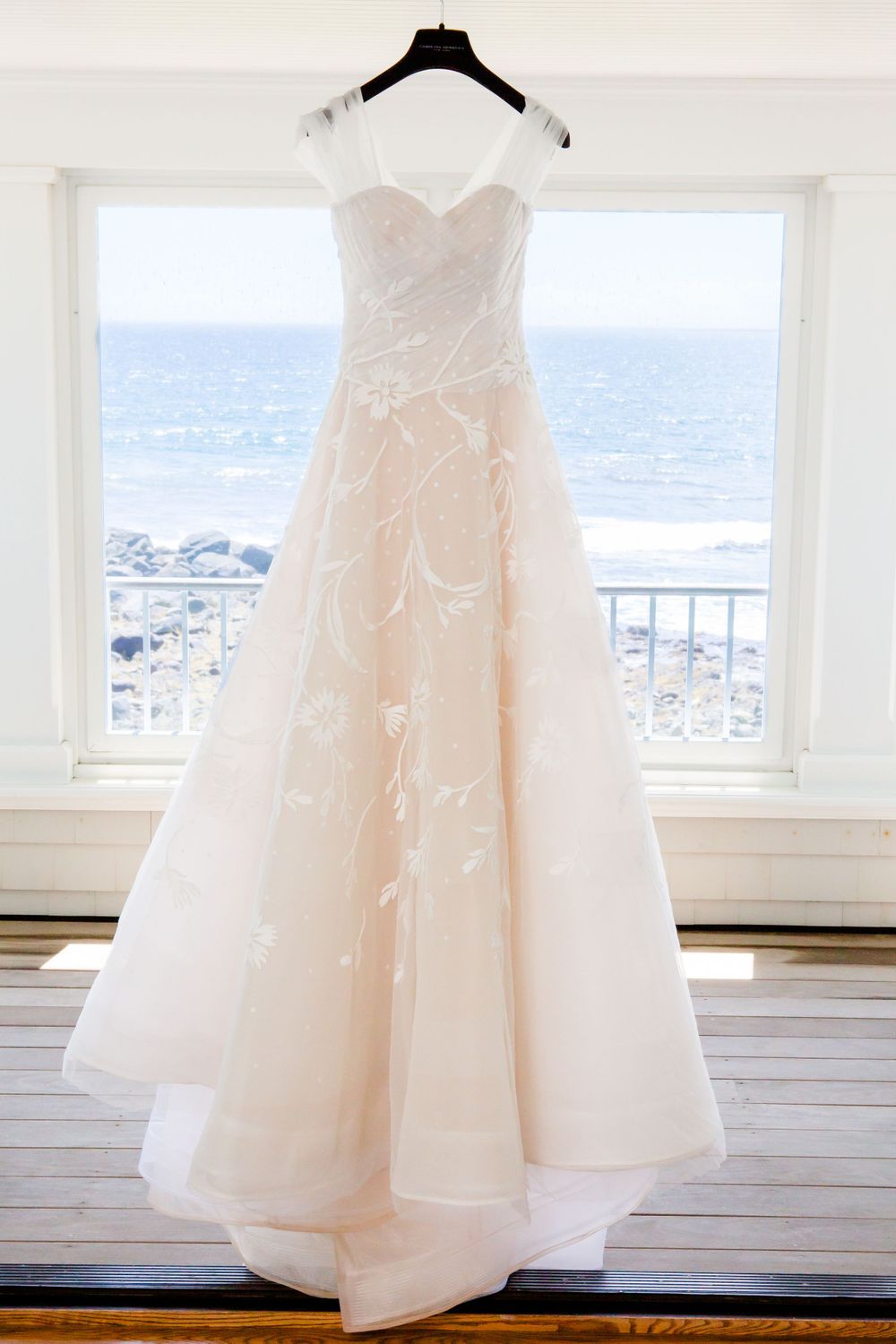 Elegant white wedding dress hanging against ocean view window.