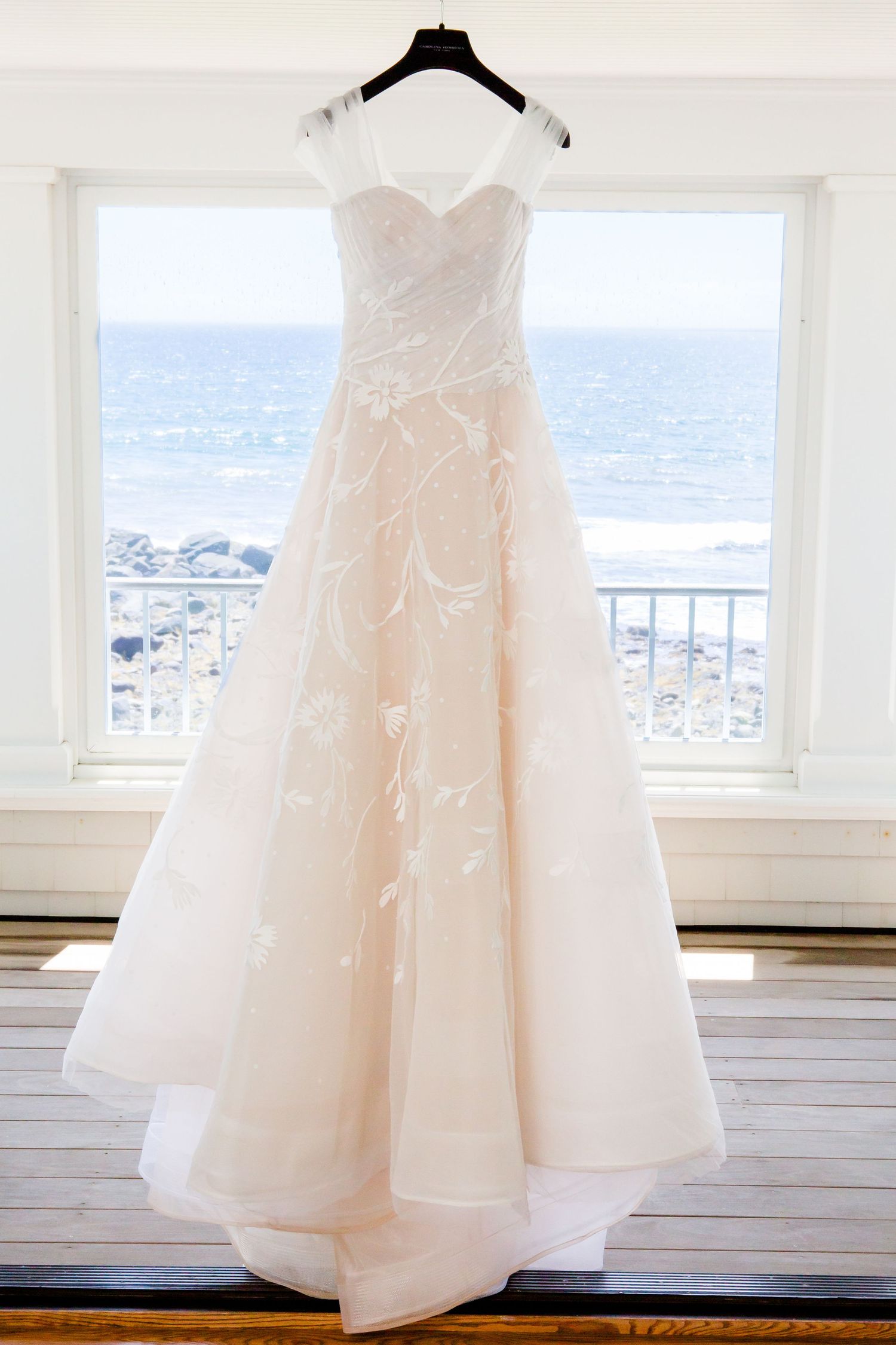 Elegant white wedding dress hanging against ocean view window.