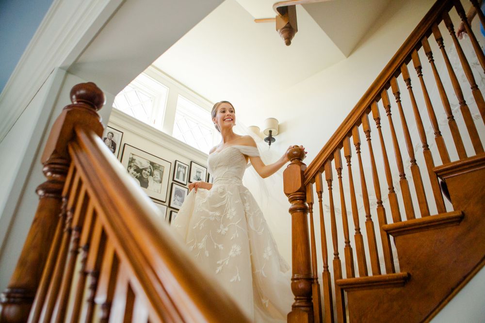 A bride in a white wedding gown descends an elegant wooden staircase.