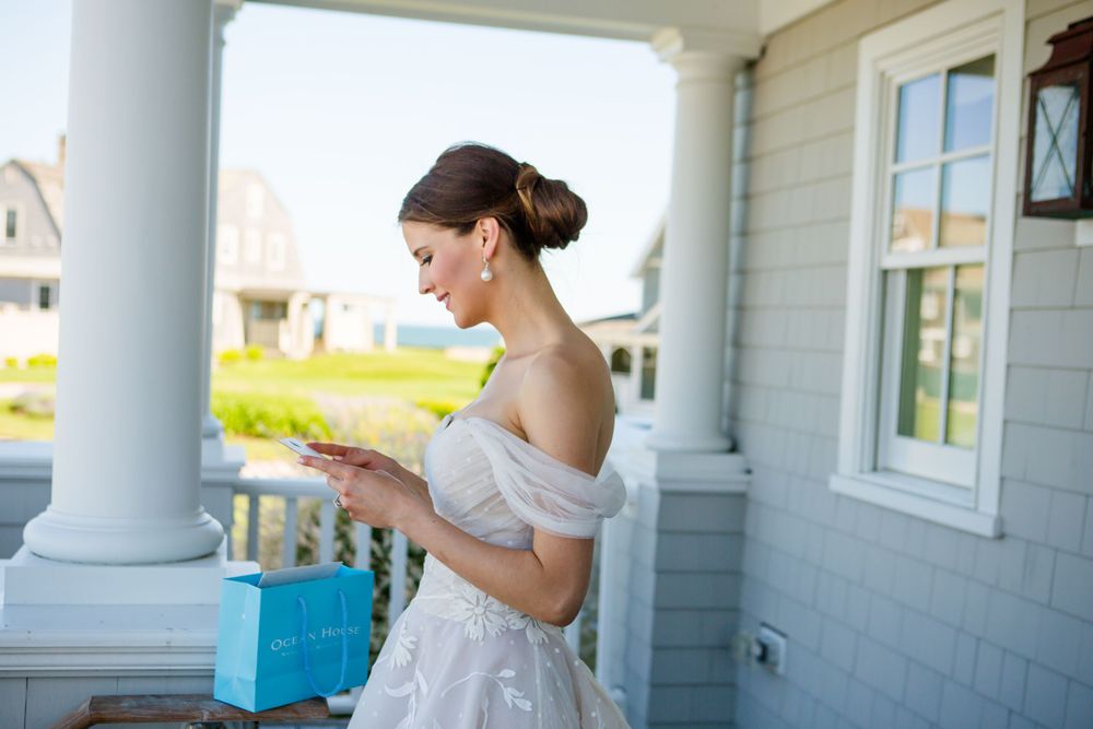 A bride in an off-shoulder gown holds a blue gift box on a white columned porch.