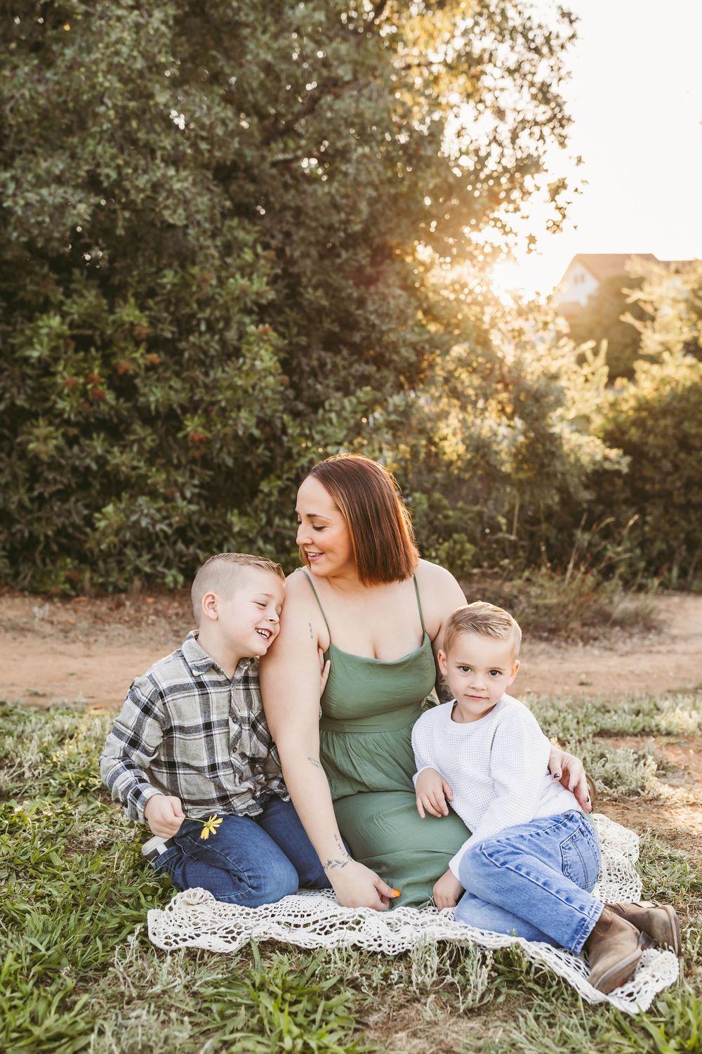 A family sits together on a blanket outdoors during golden hour as sunlight filters through trees in the background.