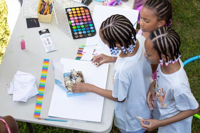 Students work together on colorful art projects at outdoor tables during a creative learning activity.