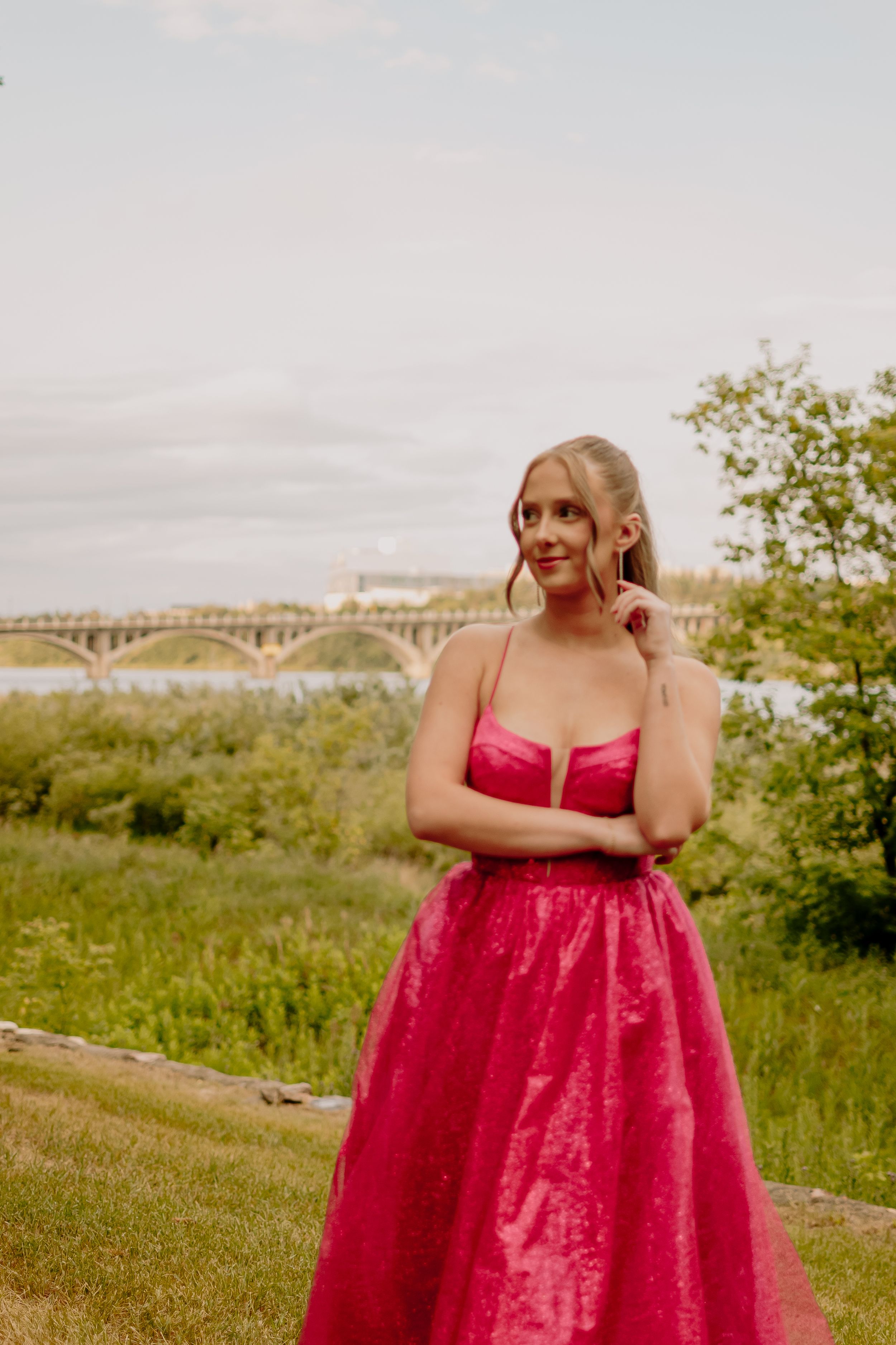 A stunning red formal gown stands out against a scenic riverside backdrop with a bridge in the distance.