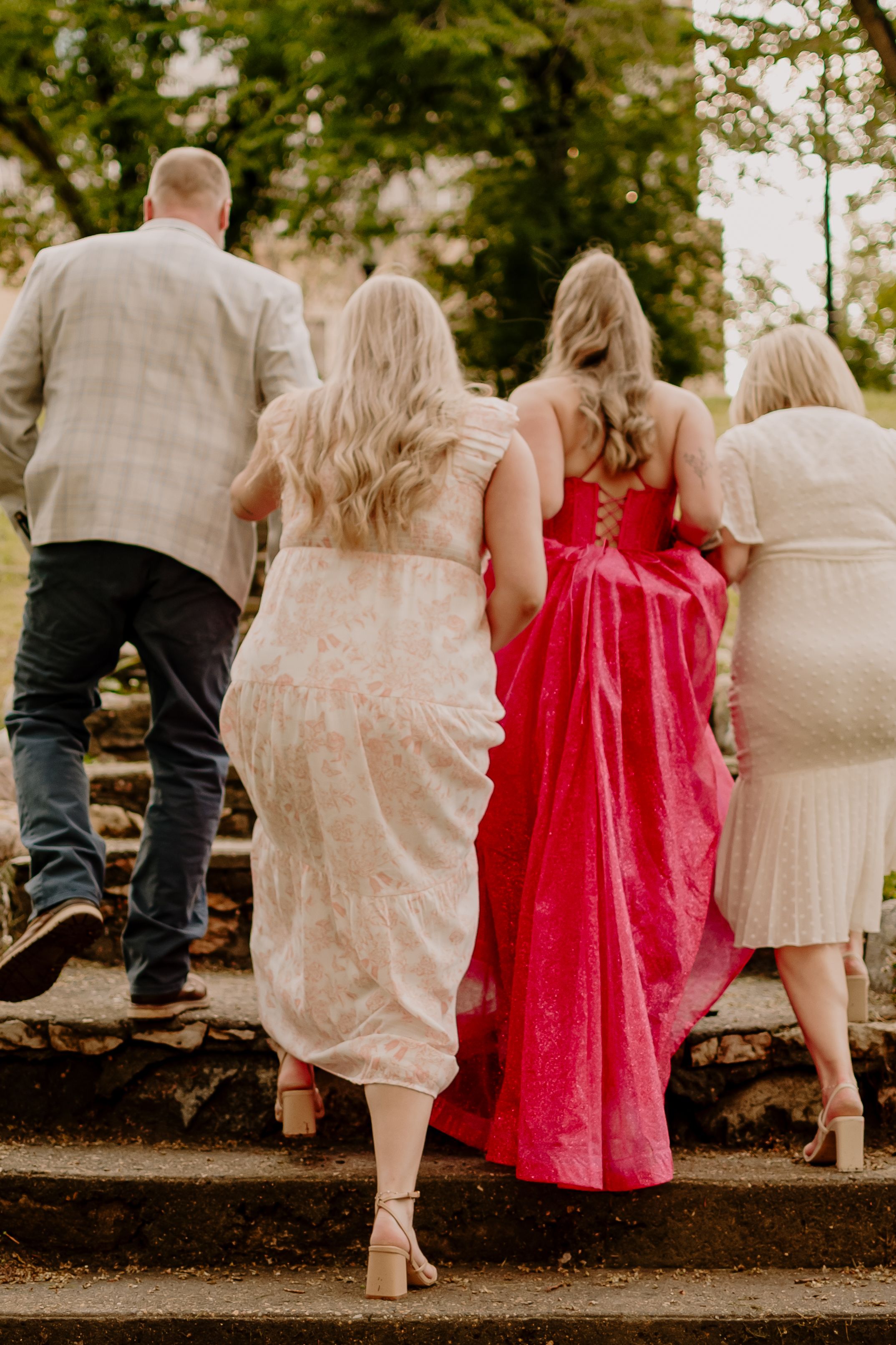 A group walking along a stone wall outdoors, with one person wearing a vibrant red dress among others in light colors.