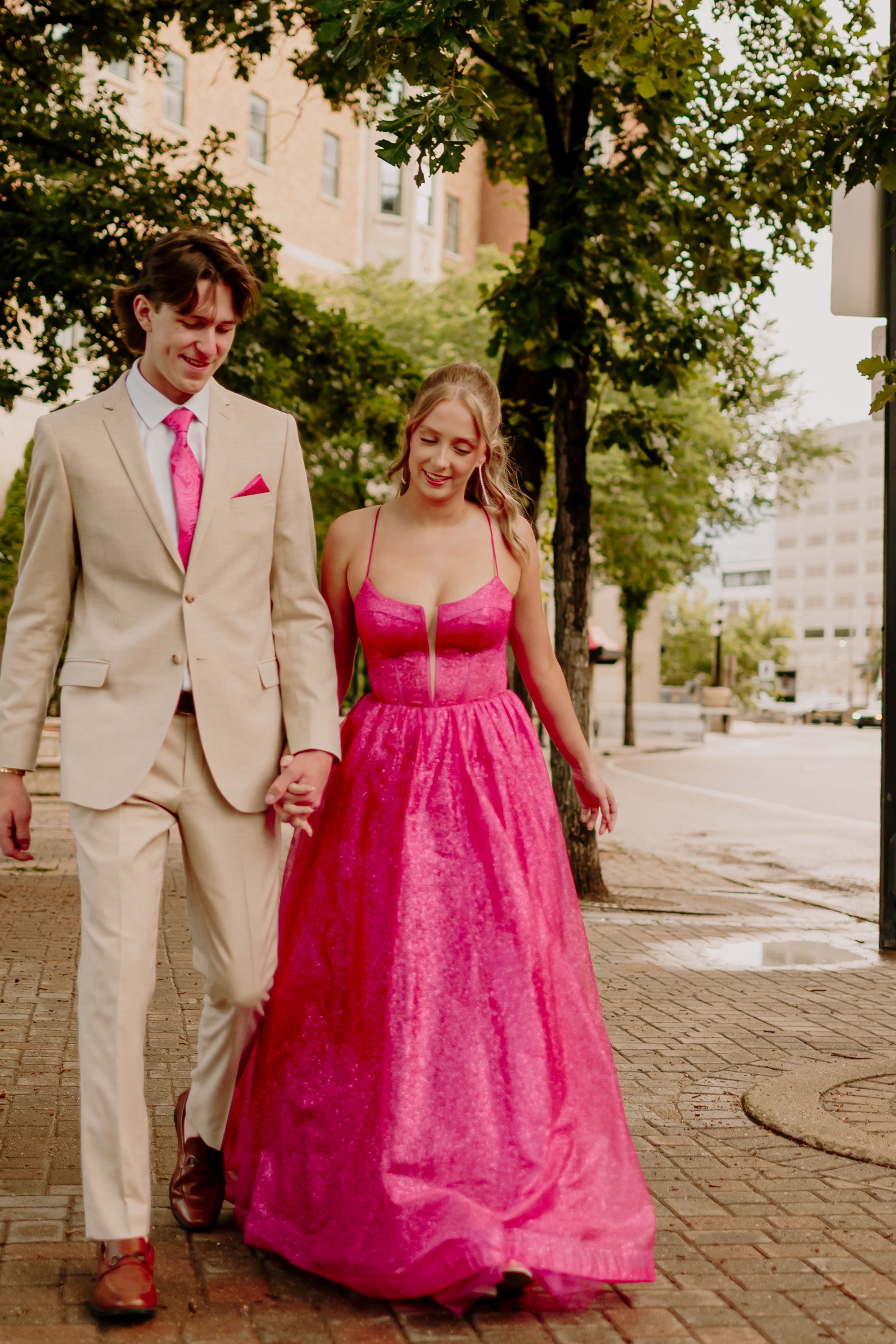 A couple walks on a cobblestone path, with the person wearing a bright pink formal dress and their partner in a tan suit.